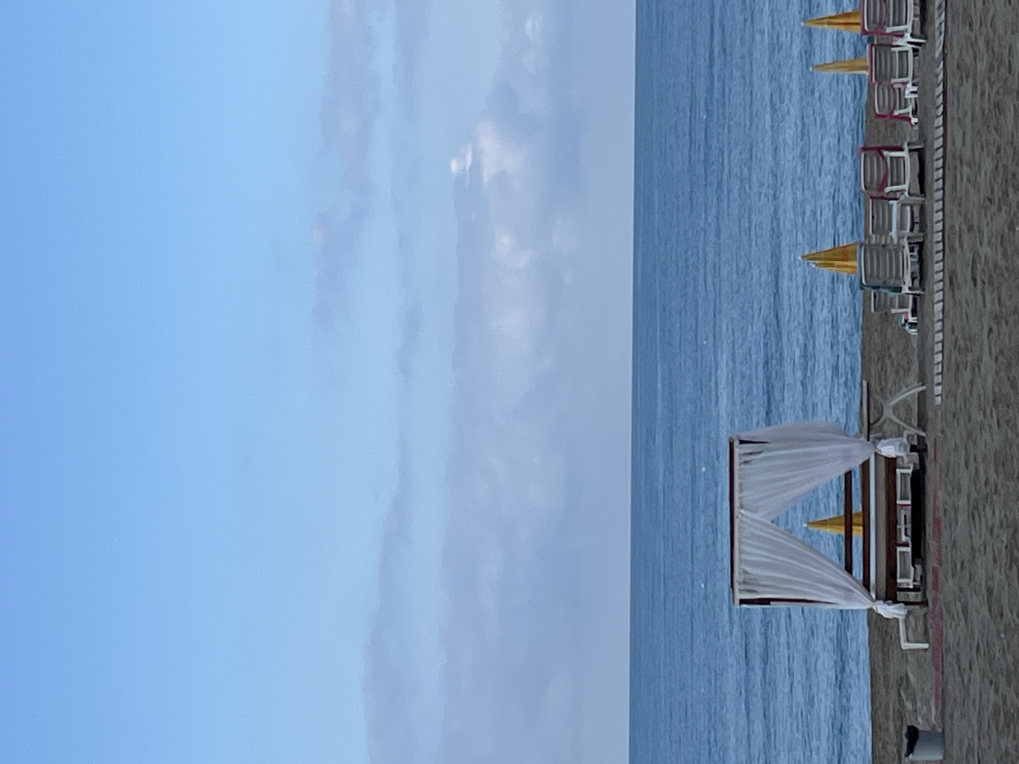 The image depicts a beach scene in Alanya, Turkey. The sky, tinged with blue and wispy clouds, occupies the left half of the frame. The right half showcases the turquoise sea meeting a sandy beach. Cabanas, complete with white flowing curtains, dot the shoreline, alongside neatly aligned rows of beach chairs with yellow and other colored sun umbrellas. There are no people visible in this image.