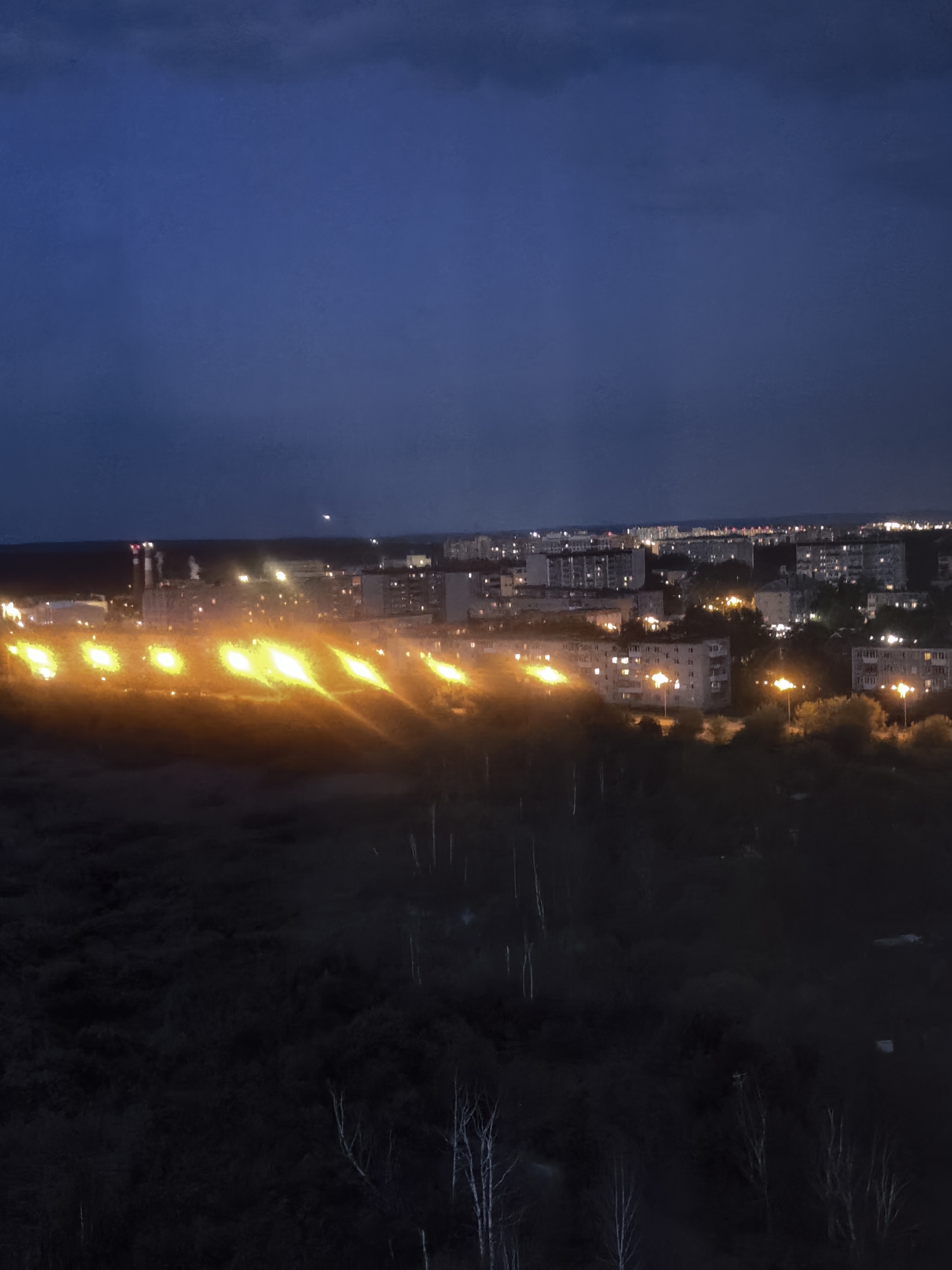 An aerial nighttime view shows a distant industrial area emitting bright orange flames or flares in the foreground, set against a dark, misty expanse of trees. Beyond the industrial zone, a city skyline is visible with numerous illuminated buildings and streetlights. The sky above is dark blue with some clouds.