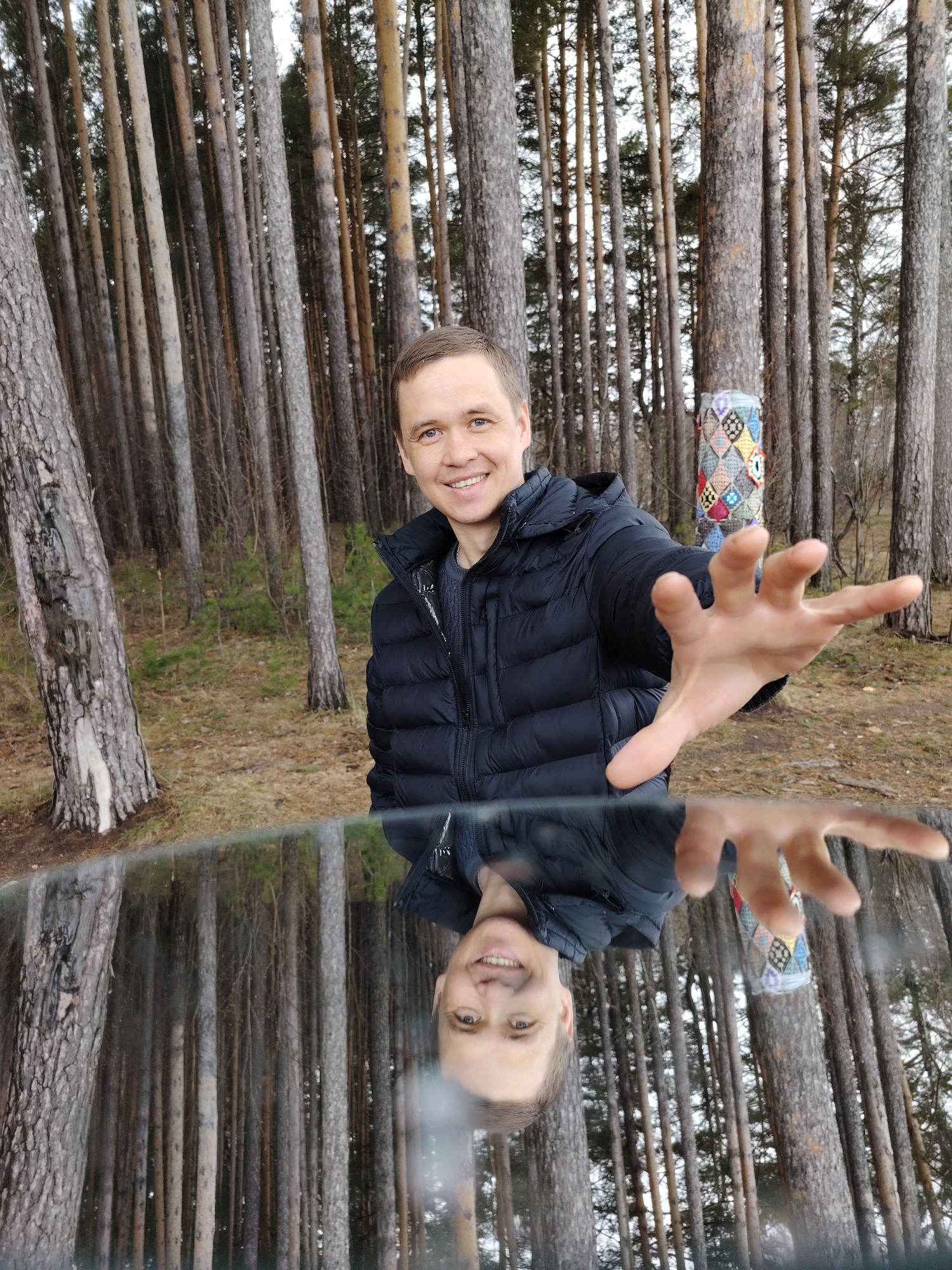 A man stands in a wooded area near Chaikovsky, Russia. He is wearing a black puffer jacket. His right arm is extended towards the camera. The reflection of the man and the forest is visible in a flat, glossy surface in the foreground. Behind the man, amidst the trees, is an object wrapped in a colorful, quilted fabric. The ground is covered with fallen leaves.