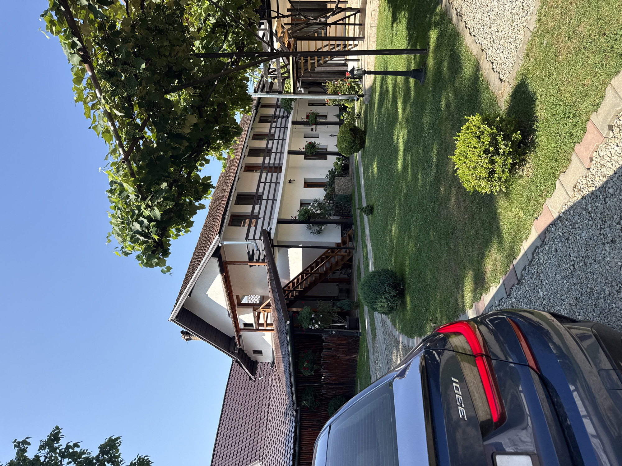 The image depicts a sunny day in Cârțișoara, Romania, in front of a white, two-story building with a dark tiled roof and wooden balconies. A dark blue car is parked on a gravel driveway to the left, with its rear visible. The building appears to be a guesthouse or residential property, with multiple windows and a wooden staircase leading to the upper floor. A well-maintained green lawn with neatly trimmed bushes surrounds the building, and a trellis with grapevines covers a portion of the outdoor area. A traditional wooden fence is visible behind the car and to the left of the building. The sky is clear blue, indicating pleasant weather. The scene conveys a sense of tranquility and rural charm.