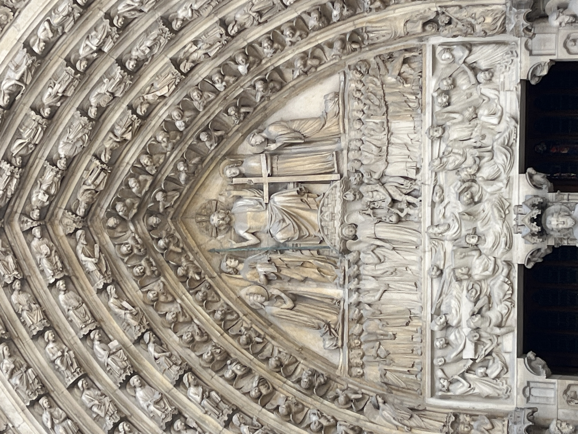 This image shows a close-up view of the tympanum and archivolts of the central portal of Notre Dame Cathedral in Paris, France. The tympanum depicts a sculpted religious scene, likely the Last Judgment or a similar biblical narrative, with numerous figures carved in stone. Above the tympanum, the archivolts are filled with rows of smaller sculpted figures, possibly saints, angels, or other religious personages, arranged in concentric arcs. The stone is weathered and light-colored, indicating it is an exterior architectural element. The details are finely carved, showcasing the artistry of medieval sculpture.