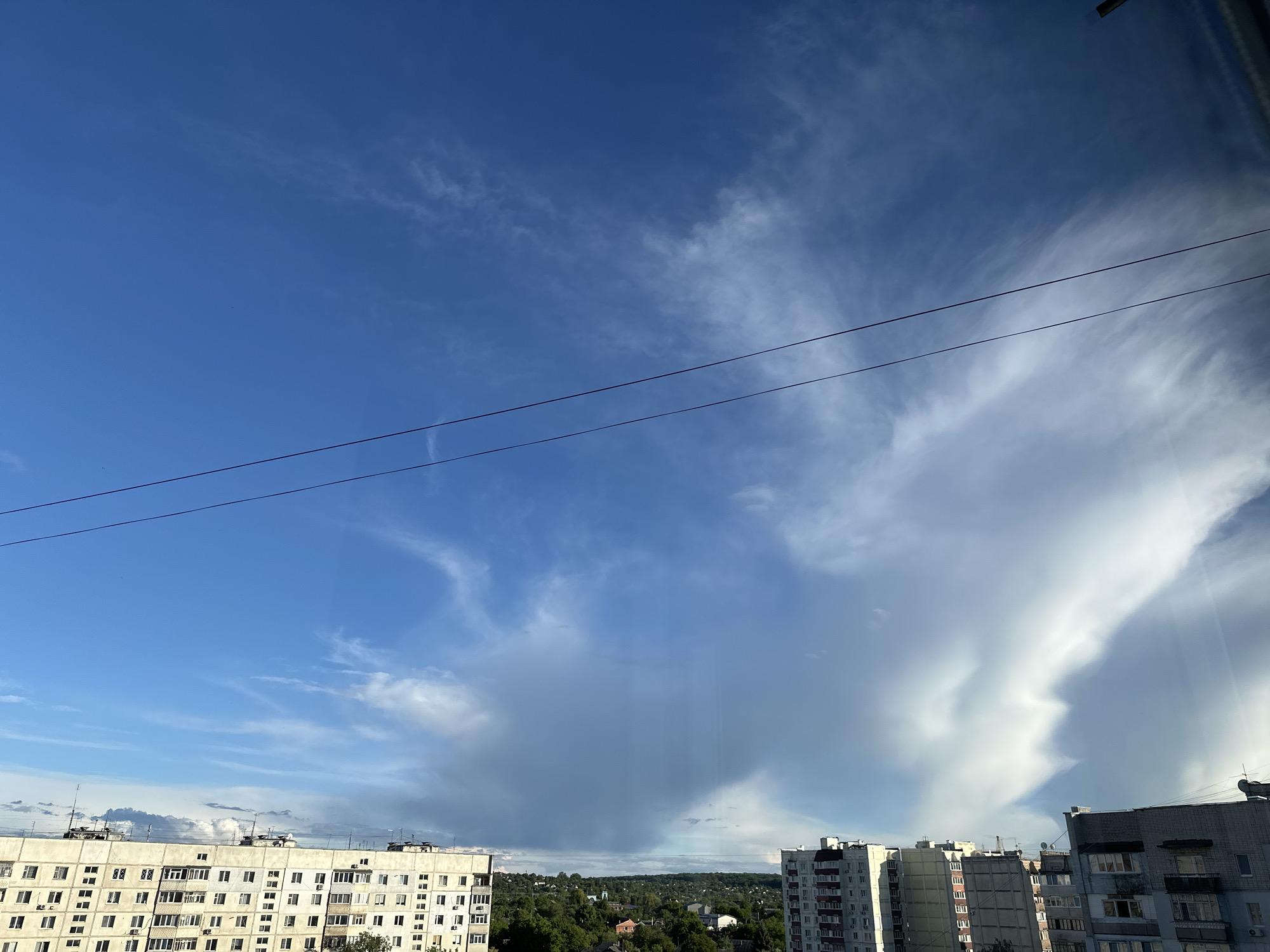 A photograph taken in the city of Unknown, Unknown, showcases a view from an elevated position, likely a window. The scene is dominated by a bright blue sky with scattered clouds of varying forms and densities. Two electrical wires stretch diagonally across the upper portion of the frame.
Below the sky, several multi-story residential buildings are visible, their facades composed of numerous windows and balconies. A line of trees is interspersed among the buildings.