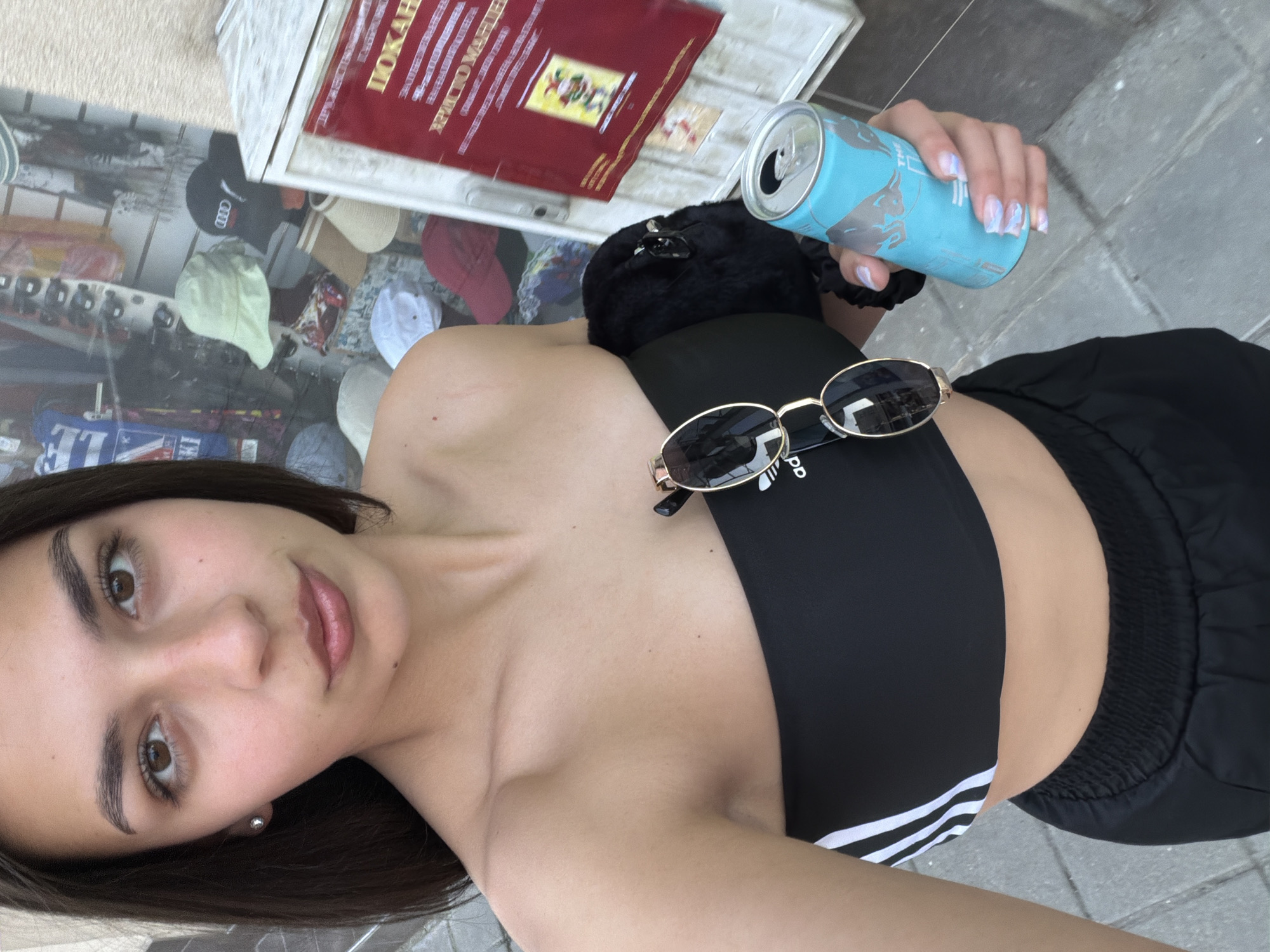 A young woman is captured in a slightly high-angle, full-frame selfie, standing on a paved sidewalk in the city of Gabrovo, Bulgaria. She has medium-length dark hair, brown eyes, and wears a black strapless crop top with white Adidas stripes on the left side, paired with black athletic-style bottoms. A pair of gold-framed aviator sunglasses hangs from the neckline of her top. In her right hand, she holds a light blue Red Bull energy drink can, and her fingernails are painted with light polish featuring a subtle design. A small, black, fluffy accessory is held under her left arm.
The background on the left features a storefront window displaying various merchandise, including a light green bucket hat, a black baseball cap with an Audi logo, and other hats and apparel. Reflections in the window show additional items, such as a shirt with a "USA" flag design. To the right, a white outdoor display cabinet or kiosk is visible, bearing a prominent red sign with Cyrillic text. Portions of the text read "ДОКАЗАН" and "ХРИСТО МАРИЕВ," accompanied by a small illustrative image, possibly a jester or cartoon figure. The setting appears to be an urban street or commercial area.