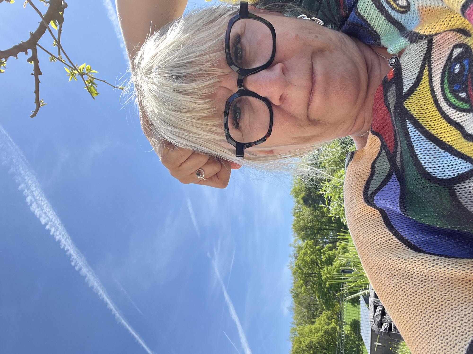 A close-up, low-angle selfie captures an older woman outdoors in Chambœuf, France, under a clear blue sky. The woman has fair skin, short, light blonde or white hair, and is wearing dark, thick-rimmed glasses that cover most of her eyes, revealing her eyelashes. Her head is tilted, and she looks directly down towards the camera, which she holds with her left hand resting behind her head, displaying a silver ring on her left ring finger. Her expression is neutral to slightly contemplative.
She is wearing a vividly colored, knitted sweater or cardigan featuring an abstract pattern with large, organic shapes in various hues including red, blue, green, yellow, and black. A delicate silver chain necklace with a small, circular pendant is visible around her neck.
The background is dominated by a bright, cloudless blue sky, streaked with several prominent white contrails from airplanes. In the upper left corner, a tree branch with fresh, light green leaves is visible, hinting at spring. The mid-ground and right side show the vibrant green canopy of numerous trees, suggesting a lush, natural outdoor setting like a garden or park. The bright sunlight indicates it is daytime, likely midday or early afternoon. A part of what appears to be dark wicker outdoor furniture is subtly visible in the lower right corner, near the green trees. There is no visible text in the image.