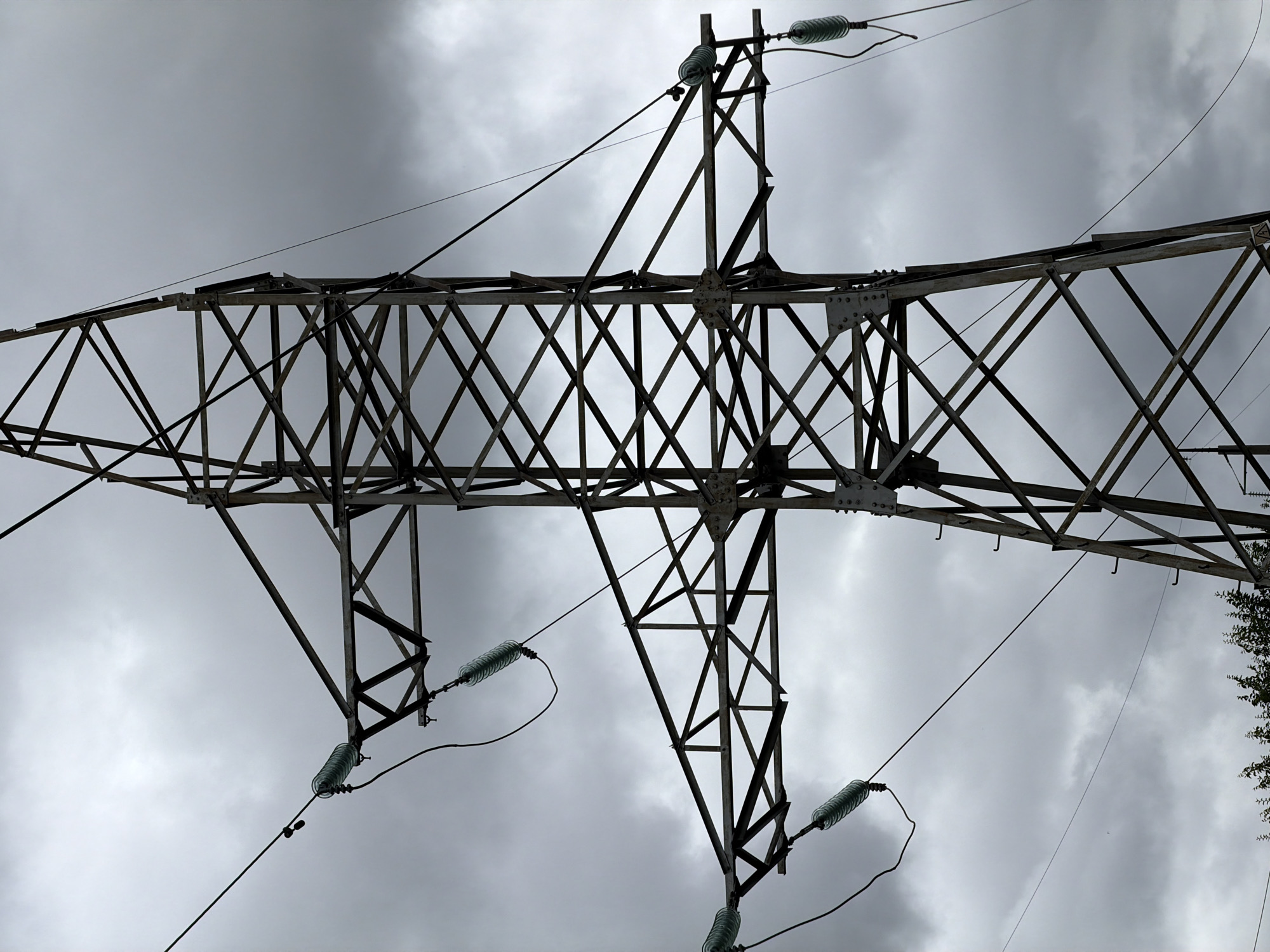 An extreme low-angle, medium shot captures the top portion of a tall, metal electrical transmission tower against a heavily overcast sky. The tower's intricate lattice structure, made of weathered metal beams, dominates the frame. Several thick, dark wires extend from insulators attached to the tower's crossbars, disappearing into the cloudy sky. The insulators, made of clear, ribbed glass, are clearly visible as they grip the wires. The sky is a uniform, pale grey, suggesting an impending storm or a generally gloomy day. The bottom of the frame shows the very tops of dark green trees, suggesting the tower is located in an area with vegetation. There are no people or any signs of recent activity visible in the image. The image appears to be taken in Ulyanovsk, Russia, based on the context provided.