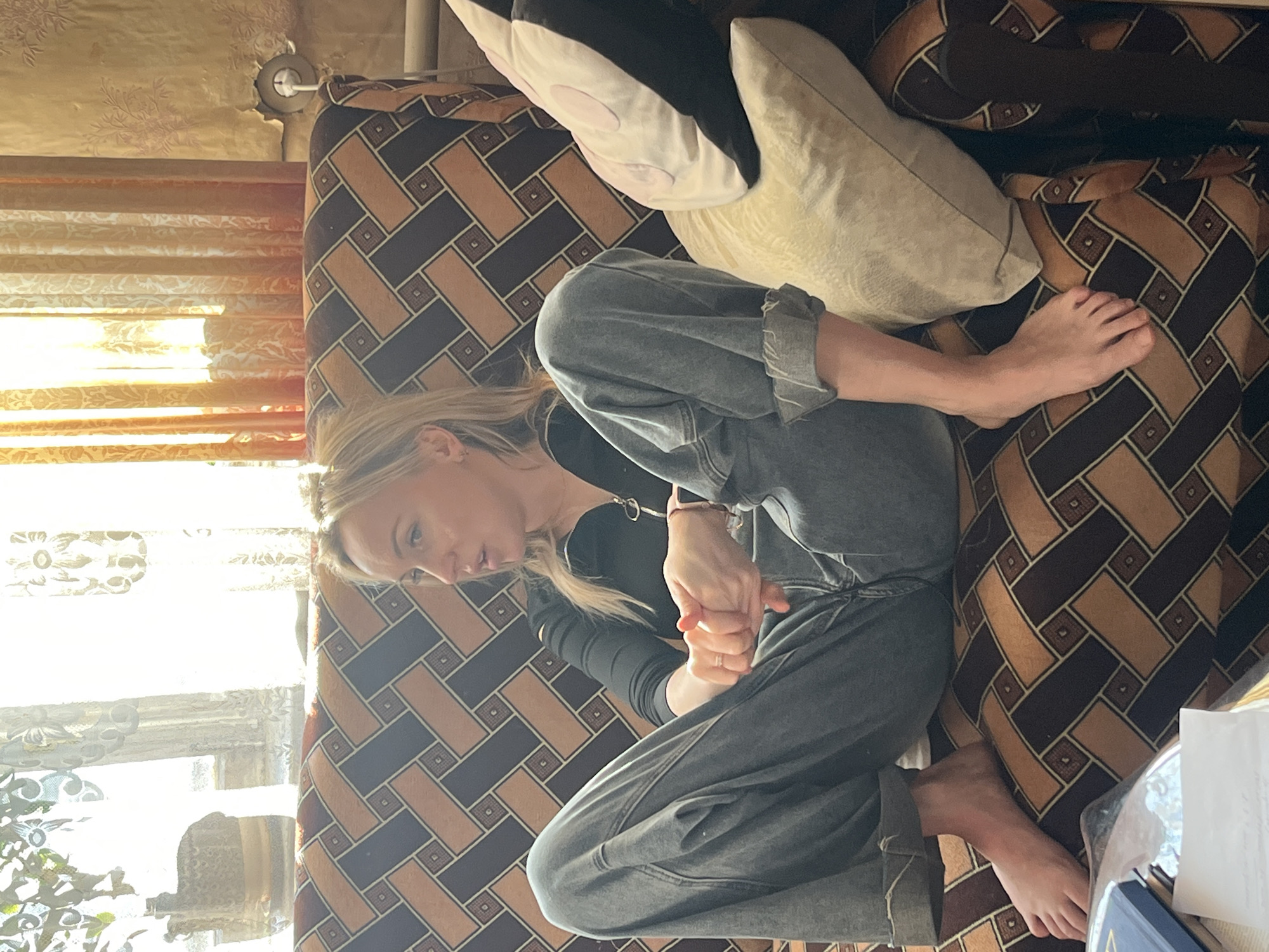 A woman is lying on a sofa. She is wearing a black top and gray pants. Her hair is blonde. The sofa has a brown geometric pattern. There are two pillows on the sofa, one white and one black and white. There is a window in the background with sunlight streaming in. The location is Podolsk, Russia.