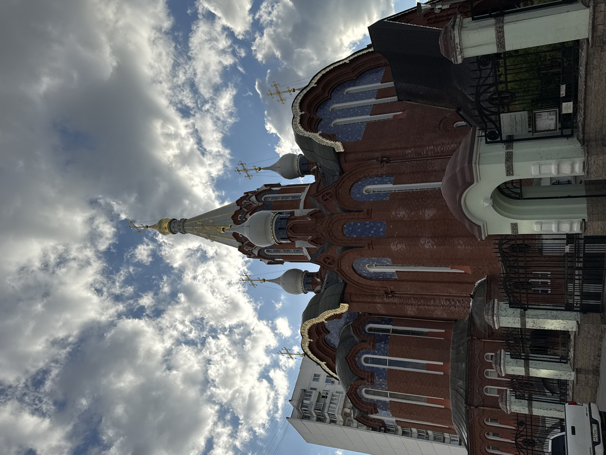 Here is a description of the image.
The image shows the Church of the Holy Trinity in Khokhly, located in Moscow, Russia. It is a brick building with multiple domes and a tall spire. The domes are decorated with blue tiles and gold trim. There are also some white accents on the building. The sky is visible in the background, and it is filled with white clouds. Parts of buildings are also visible at the bottom of the photo.