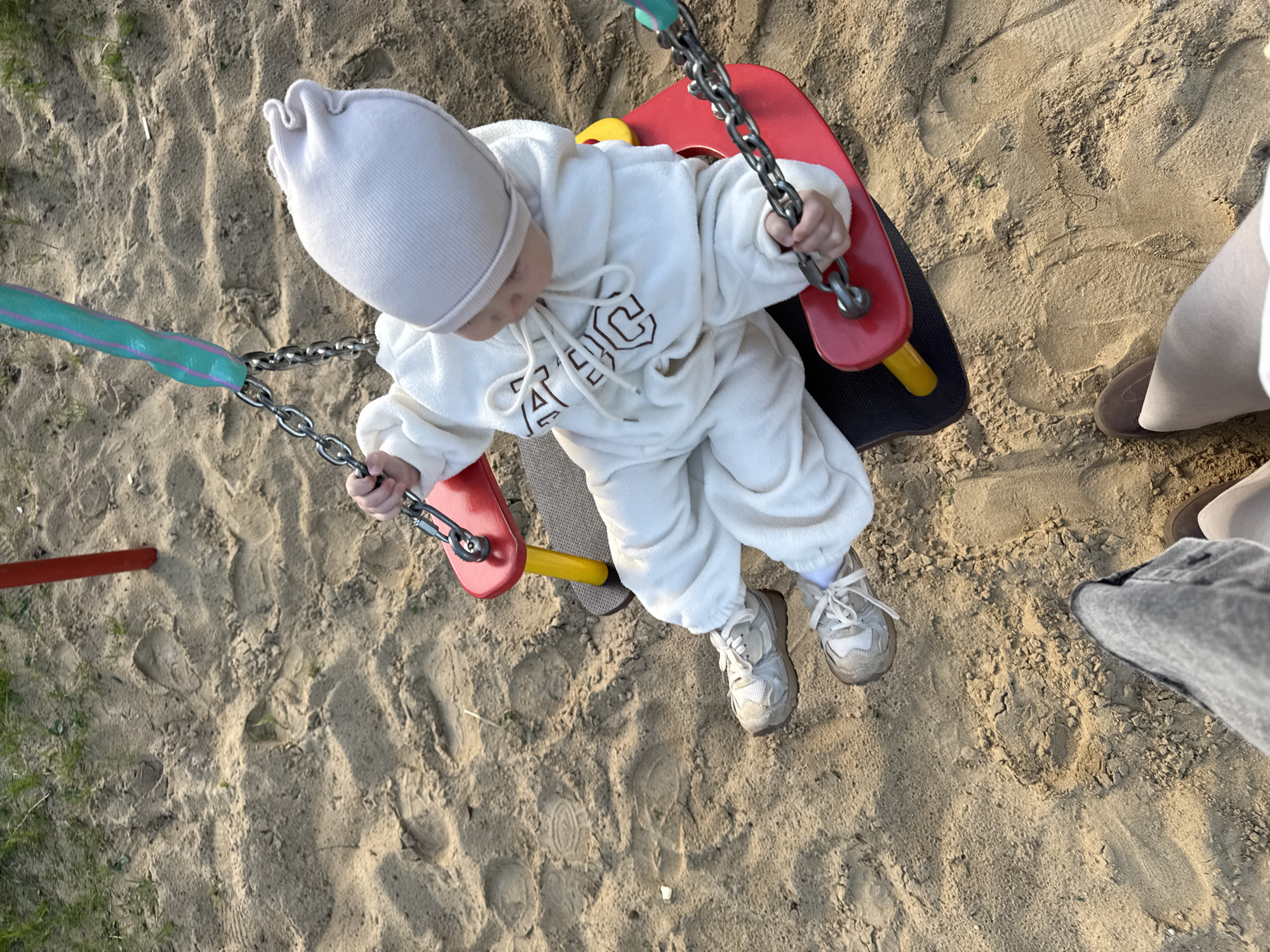A toddler, wearing a white hooded jumpsuit and a matching hat, sits on a red and black swing set in a sandbox. The swing is suspended by metal chains with teal-colored grips. The toddler is holding onto the chains with both hands. The sandbox is filled with light brown sand. Two adult feet are visible to the right of the toddler, one wearing beige shoes and the other wearing gray denim. The location is a playground setting in Vladimir, Russia.