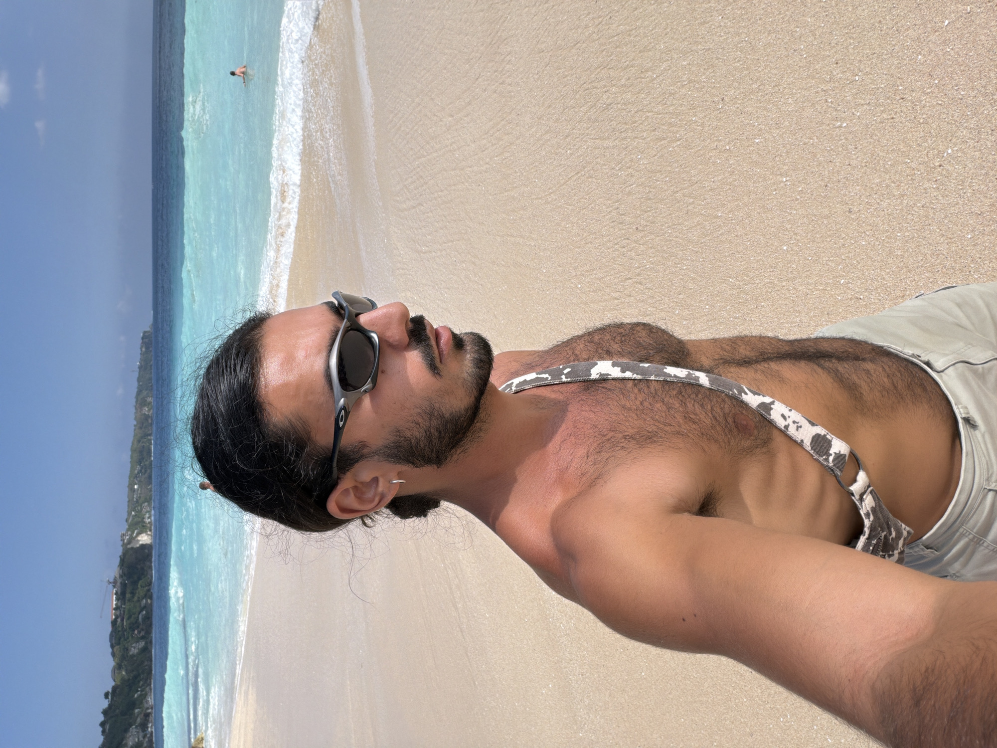 In Kuta Selatan, Indonesia, a man with dark hair and a beard, wearing sunglasses, a patterned strap, and light-colored shorts, poses for a selfie on a sandy beach. The ocean, with its clear turquoise water and white-capped waves, stretches out behind him, meeting a deep blue sea under a bright, cloudless sky. In the background, a cliffside overlooks the coastline, with a small structure visible on its edge. A person is swimming in the water in the distance.