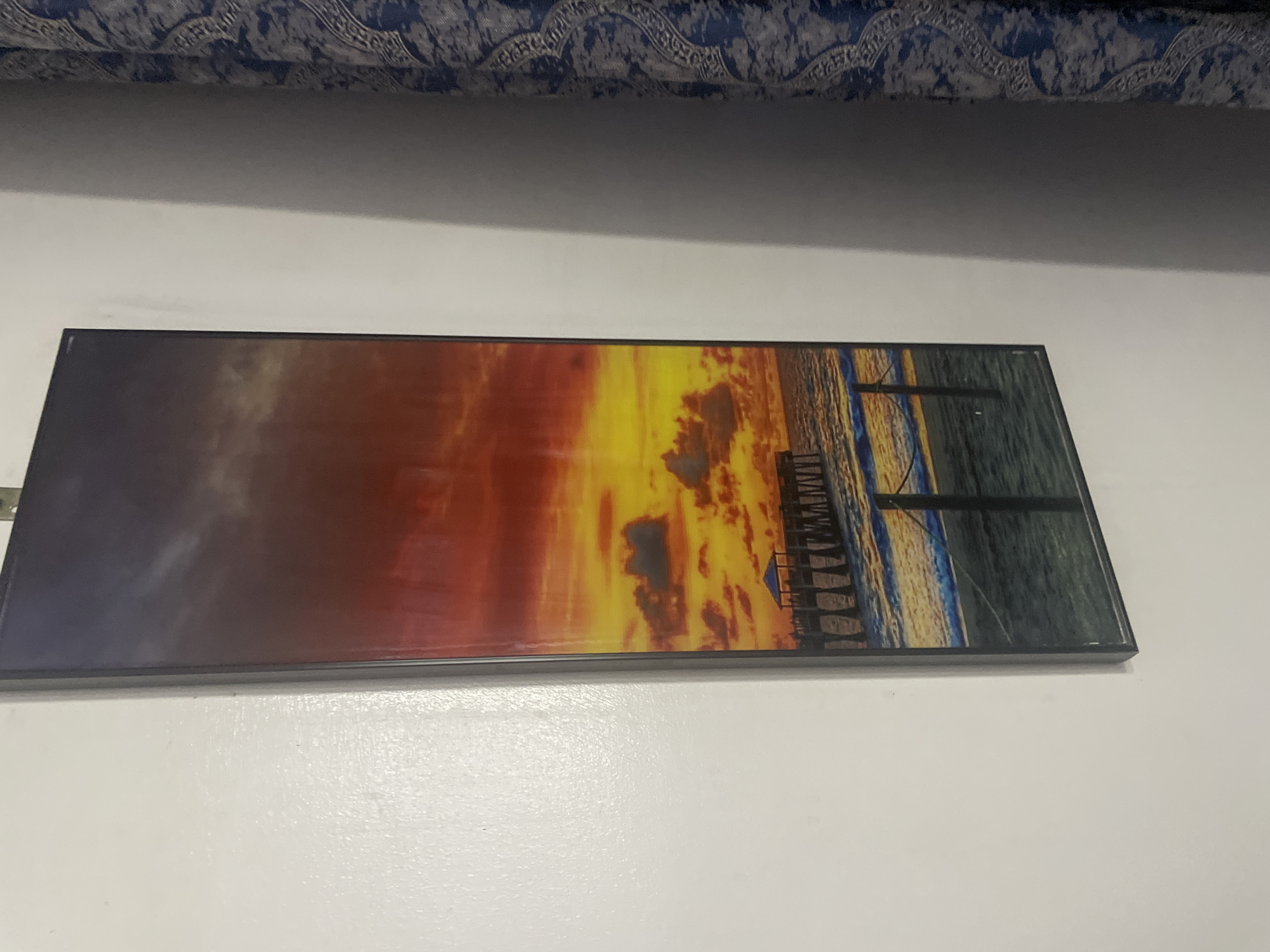 A landscape painting hangs on a white wall, with a blue patterned textile visible above. The painting depicts a sunset over the ocean, with a pier extending into the water. The colors are primarily orange, yellow, and red. A black frame surrounds the painting. The location of the scene is unknown and cannot be assumed to be Abuja, Nigeria.