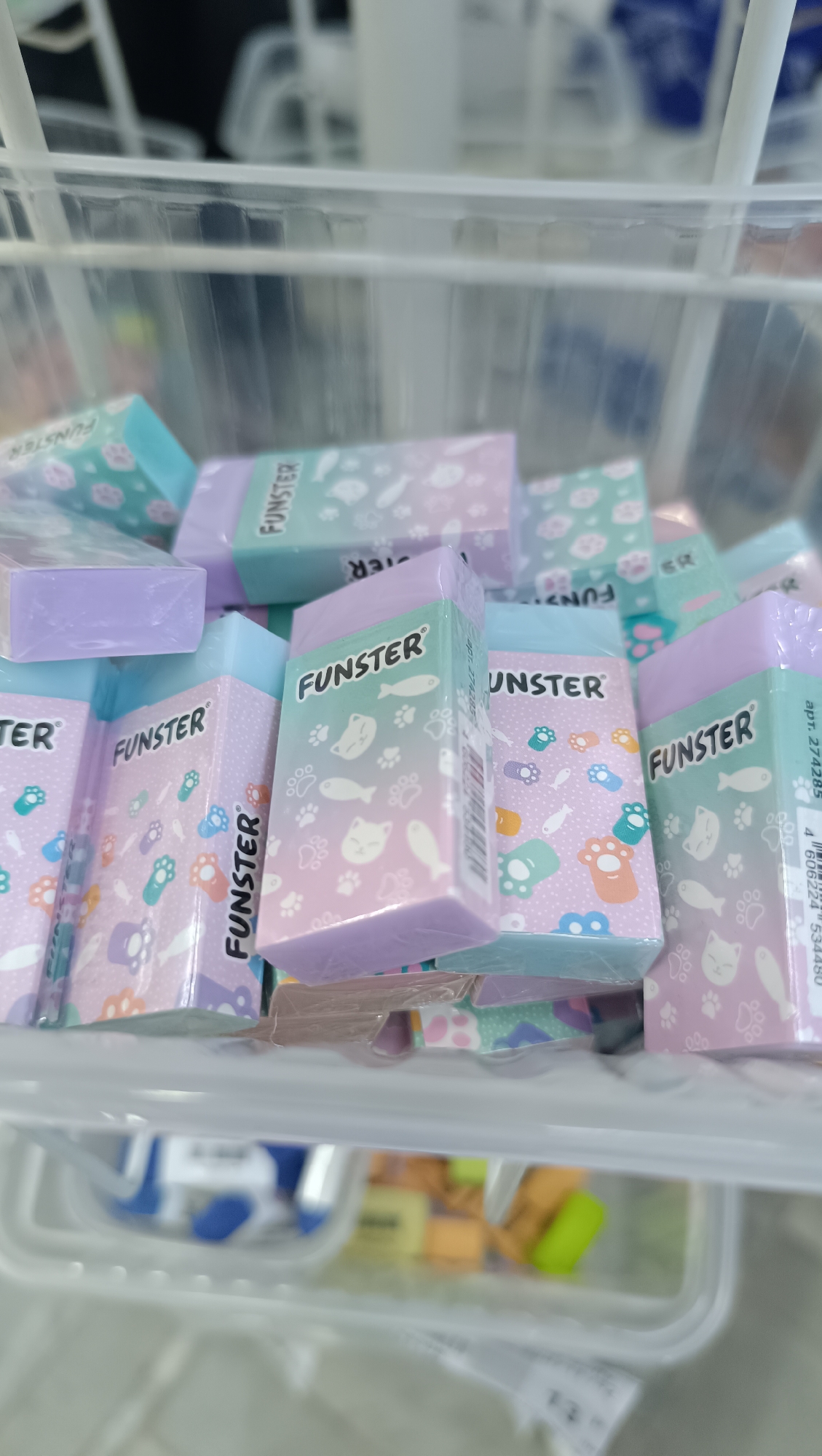 The image displays a close-up, eye-level view of a white plastic bin filled with a quantity of pastel-colored rectangular erasers. The erasers are decorated with various cartoonish animal paw prints and fish designs in shades of pink, blue, and purple. Some of the erasers have "FUNSTER" printed on them. The bin appears to be made of white plastic with a grid-like pattern. The background is blurred, showing more white plastic containers and some out-of-focus blue and white objects. The lighting suggests an indoor retail or storage environment. There are no people or discernible human interaction visible in the image. The scene is consistent with a display of stationery or art supplies. There is no information within the image to suggest a location in Chaikovsky, Russia.