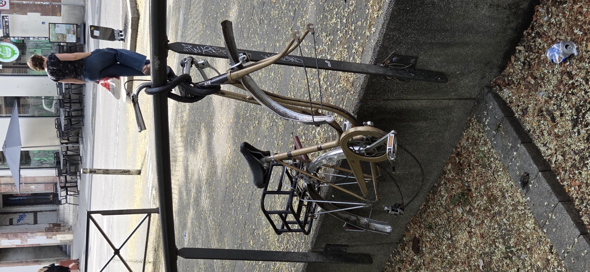 A vintage, tan-colored bicycle with a black seat and a black rear rack is secured to a black metal railing with a thick black cable lock. The bicycle is leaning against a concrete wall, with its front wheel off the ground. The scene is outdoors, on a street in Strasbourg, France. Scattered on the ground are fallen leaves and a crumpled aluminum can. In the background, there are tables and chairs set up outside a building with an umbrella providing shade. A woman in blue pants and a dark patterned shirt is walking away from the camera.