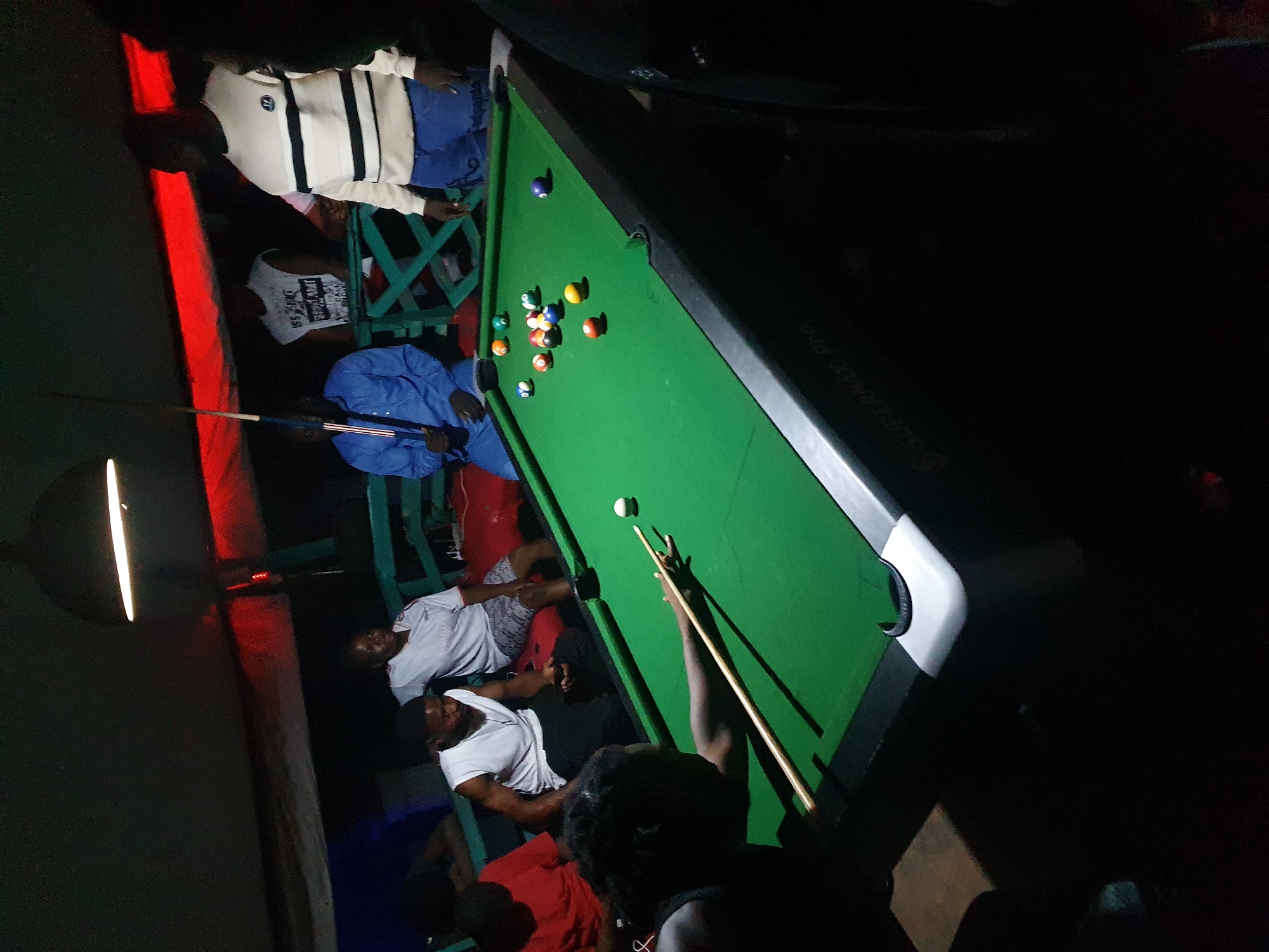 A low-lit indoor scene depicts a group of people gathered around a pool table in what appears to be a bar or recreational area. The green pool table is partially set with billiards balls, and a person is actively lining up a shot with a cue stick. Several individuals are seated or standing around the table, seemingly observing the game. The room is dimly lit, with some red lighting in the background, which gives the scene a slightly hazy atmosphere. The environment appears casual and informal, suggesting a leisure activity or social gathering. The location is assumed to be in Lokoja, Nigeria.