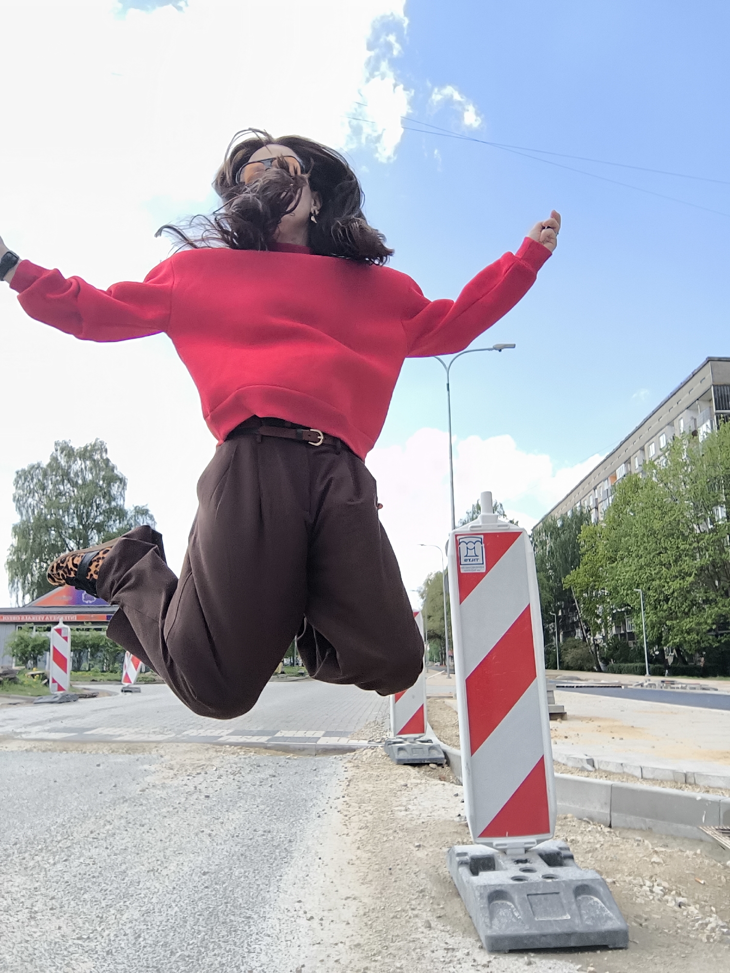 A woman is captured mid-air, jumping in front of what appears to be a construction zone in Riga, Latvia. She is wearing a red sweater, brown wide-legged pants with a brown belt, and animal print shoes. The background reveals elements of the urban landscape, including a building, trees, streetlights, and red-and-white striped traffic barriers. The ground in the foreground has a gravelly appearance, indicating the road work in progress. The sky is blue with scattered clouds.