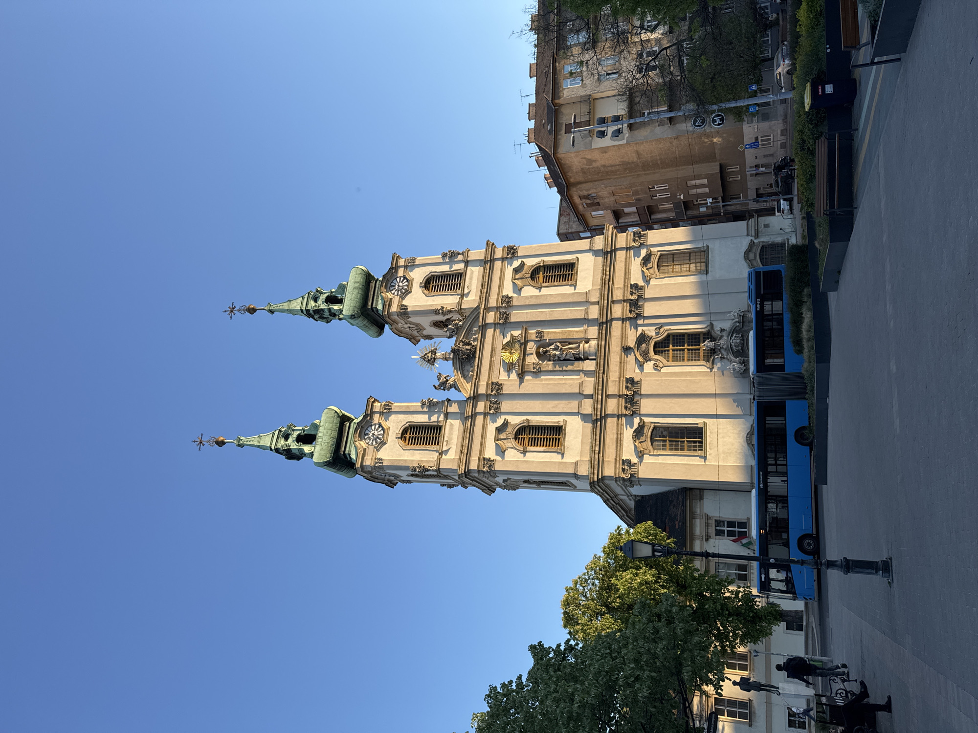 This photograph shows the Church of St. Anne in Budapest, Hungary. The church features two tall, ornate towers with green spires against a clear blue sky. The facade of the church is detailed with statues and windows. Adjacent to the church is a modern building and a blue bus parked on the street. Trees and benches are near the bus, and pedestrians walk on the sidewalk. The lighting suggests it is either early morning or late afternoon.
