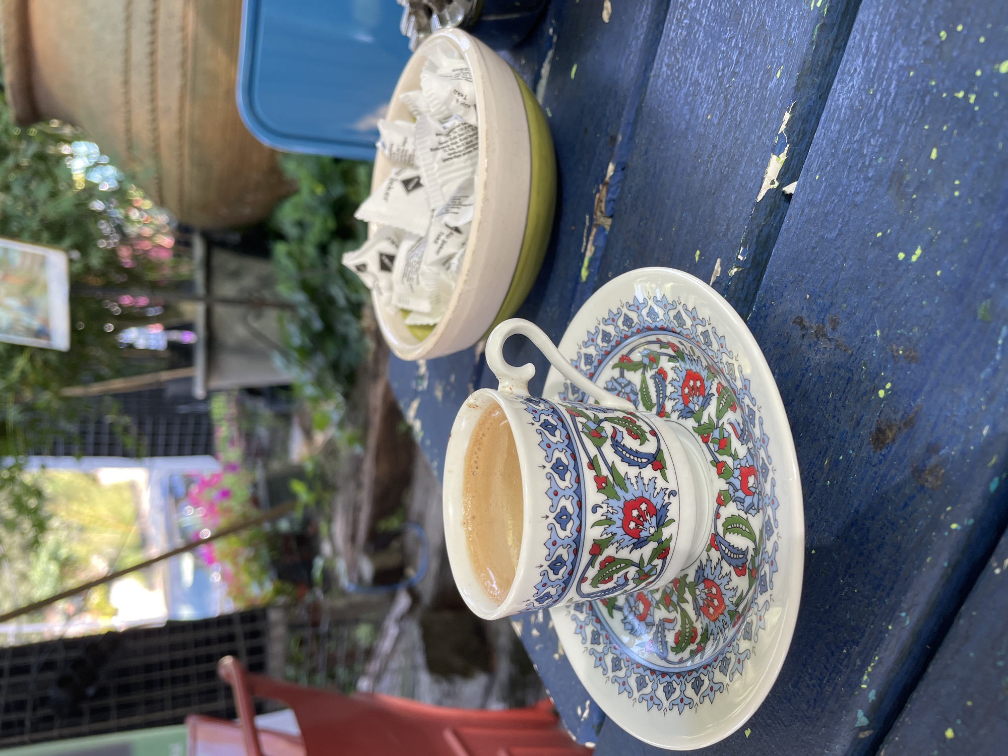 A close-up shot captures a cup of Turkish coffee, adorned with a traditional blue and red floral pattern, resting on its matching saucer. The set sits on a weathered, dark blue wooden table, its surface showing signs of wear and paint chips. To the right of the coffee cup, a shallow bowl overflows with individually wrapped sugar packets. In the background, out of focus, greenery suggests an outdoor setting, possibly a garden or courtyard. The lighting is natural, indicating daylight. The scene evokes a tranquil, relaxed atmosphere, likely in a cafe or a residential garden in Üsküdar, Turkey, given the cultural context of Turkish coffee and its typical serving ware.
