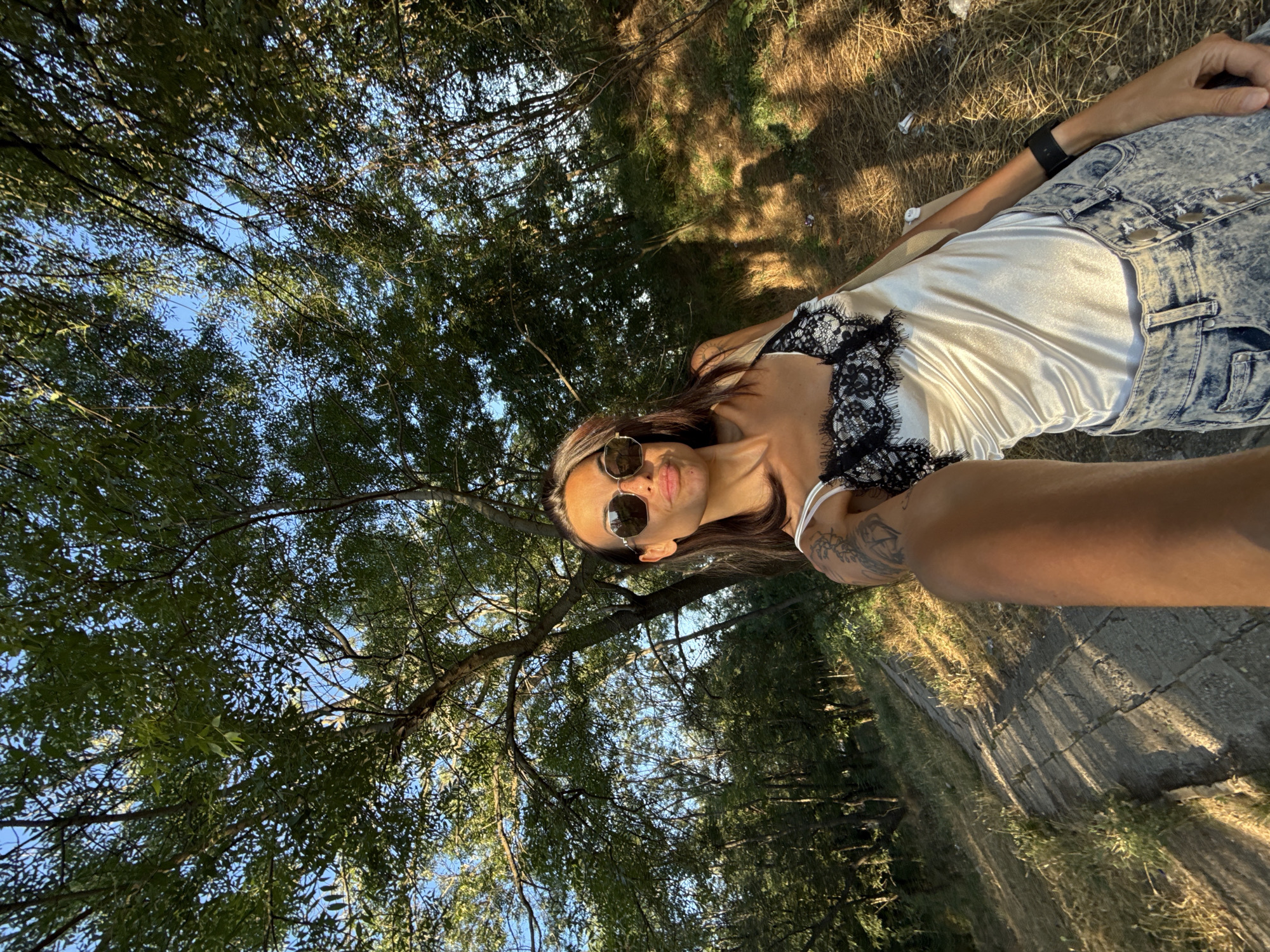 A woman is taking a selfie in a park in Pleven, Bulgaria. She is wearing a white satin camisole with black lace trim and acid-wash jeans. She has sunglasses on and her arm is extended, holding the camera. The background is a wooded area with trees and a path made of stone slabs. The sun is shining, casting shadows on the path. There are some scattered pieces of trash on the ground.