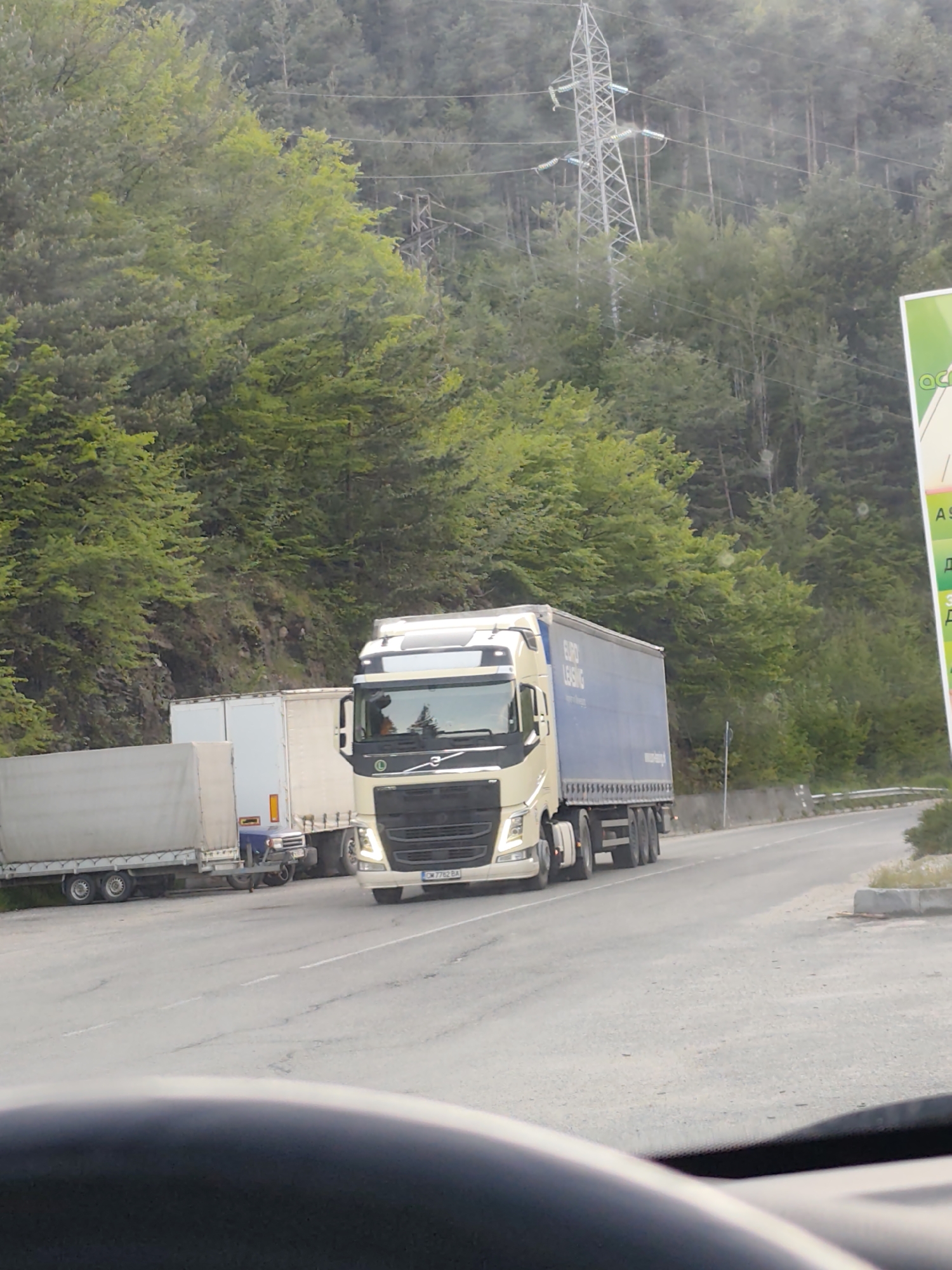 The image shows a road in Hasovitsa, Bulgaria, with two trucks. The first truck is a Volvo semi-trailer truck with a beige cab and a blue trailer, bearing the logo "EURO LEASING". It is moving on the road. Next to the Volvo is another semi-trailer truck parked on the side of the road. The background features a forested hill, a power transmission tower, and a sign on the right side of the frame, partially obscuring some text. The photograph is taken from inside a vehicle, as the dashboard is visible in the foreground.