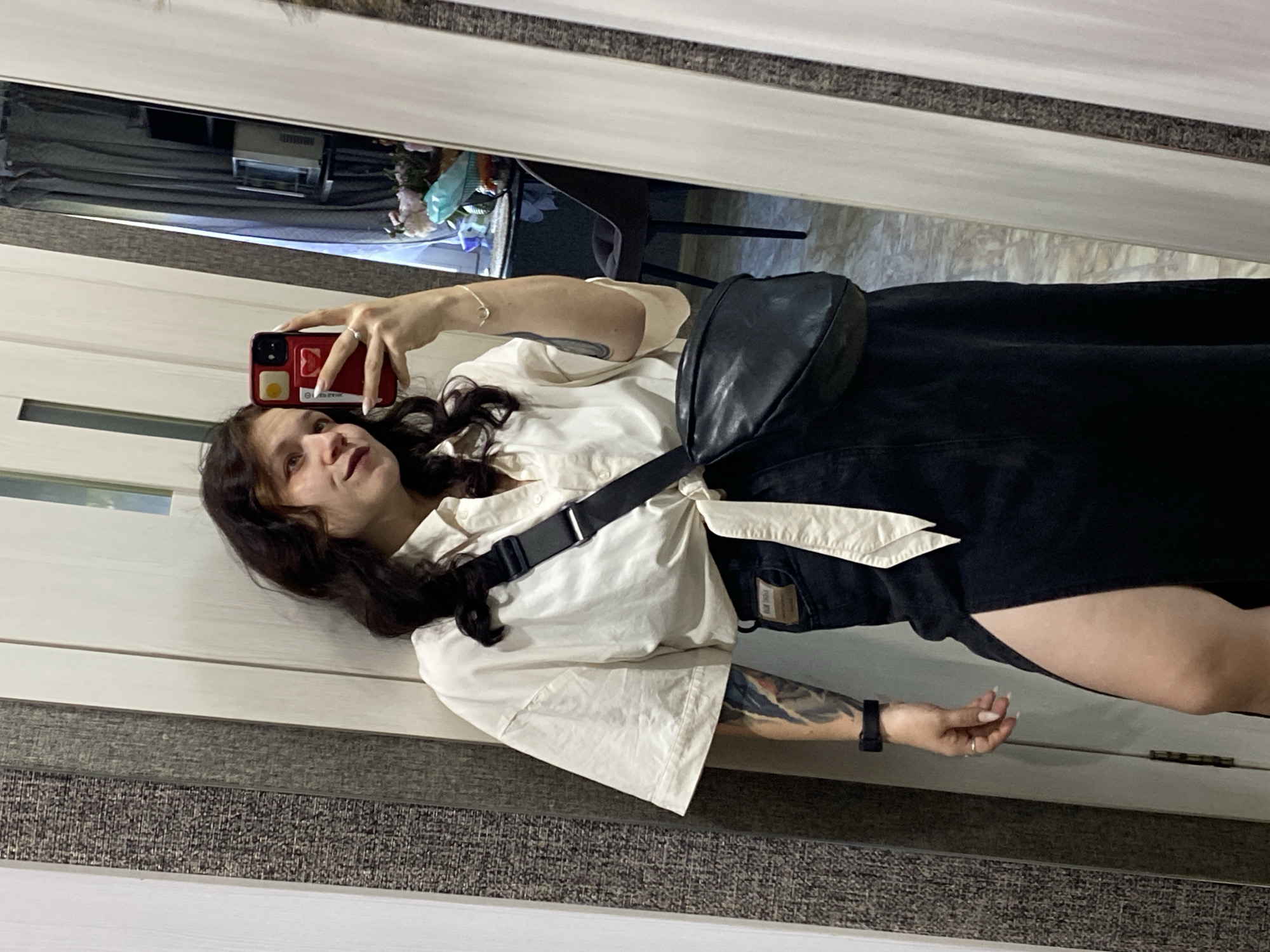 A young woman with dark, curly hair takes a mirror selfie in what appears to be a residential hallway. She is wearing a cream-colored, oversized collared shirt tied at the waist, a black denim maxi skirt with a high slit, and a black leather crossbody bag. She has tattoos on her left arm and wears a black watch on her left wrist and rings on her fingers. She is holding a red smartphone. The environment is a hallway with textured wallpaper and light-colored doors. In the mirror's reflection, a chair and some items on a shelf are visible, suggesting the presence of a room beyond the hallway. Based on available information about the provided image, it is highly probable that this scene is located in Chaikovsky, Russia.