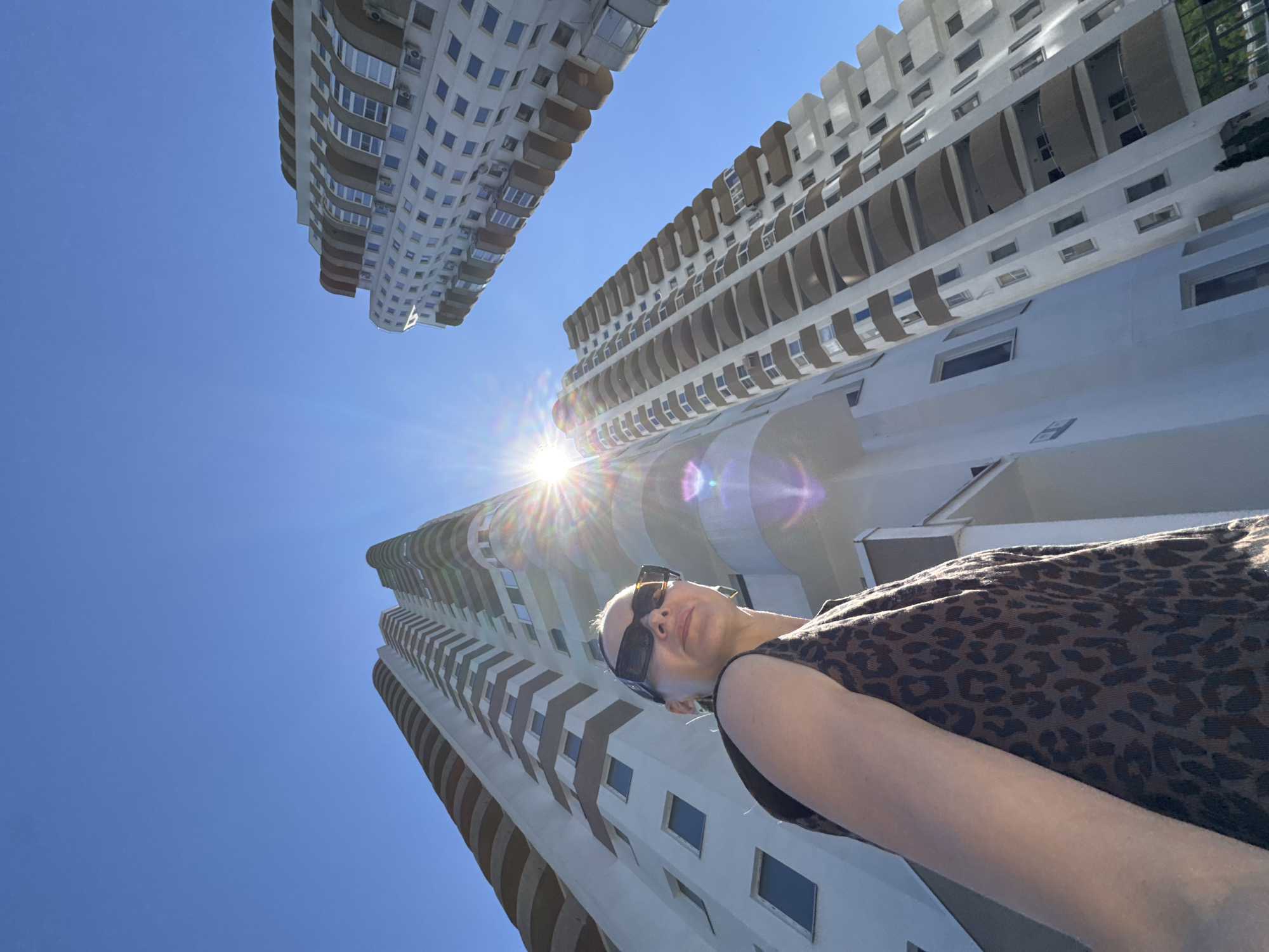 A low-angle shot captures a woman standing outdoors with two tall apartment buildings in the background against a clear blue sky. The woman is positioned in the foreground on the left side of the frame, looking upwards and slightly towards the viewer. She has dark hair pulled back and is wearing large, dark sunglasses and a sleeveless, brown, animal-print top.
The two apartment buildings are modern, white structures with rounded balconies and numerous windows. The building on the left is taller and closer to the viewer, while the one on the right is slightly further away and angled towards the left. The sun is bright and positioned between the two buildings, creating a lens flare with visible rays and light streaks. The lighting suggests it is daytime and sunny. The environment appears to be an urban setting, likely a residential area, in Sochi, Russia, given the architectural style and clear, bright weather often associated with the city.