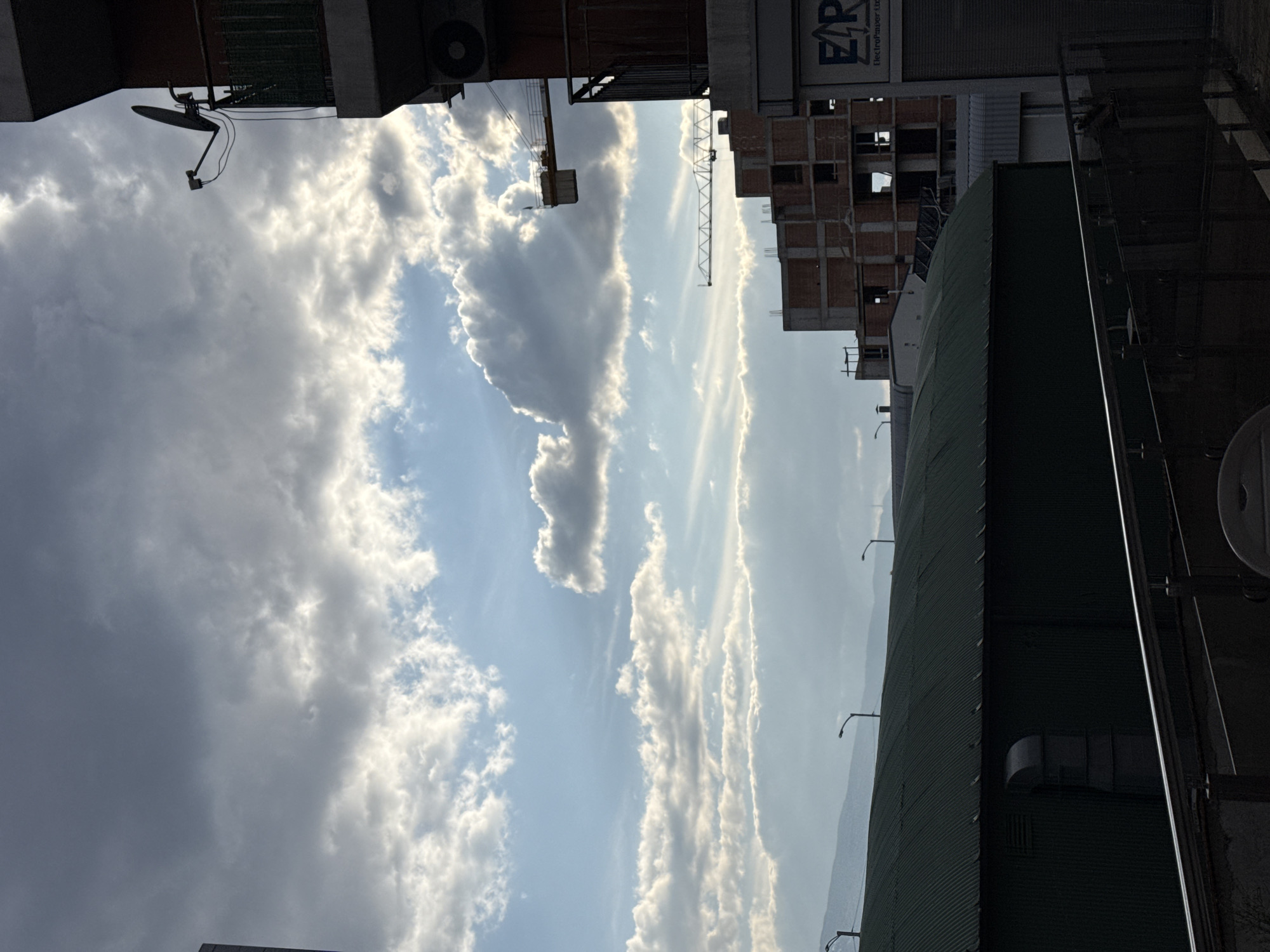 The image depicts an urban landscape in Sofia, Bulgaria, characterized by a mix of residential structures, industrial-type buildings, and ongoing construction under a dynamic, partly cloudy sky. The lighting suggests either early morning or late afternoon, with sunlight filtering through and illuminating the edges of the clouds.
In the immediate foreground, to the right, is a section of a multi-story building featuring a balcony from which a black satellite dish extends. Below the balcony, an air conditioning unit is affixed to the building's facade. The building material appears to be a reddish hue.
Stretching across the lower left and central portion of the midground is a large, dark green building with a corrugated metal roof, possibly a warehouse or industrial facility. A prominent light gray exhaust vent is visible on its side. Multiple streetlights are silhouetted against the sky above this building.
Further into the midground and to the right, several unfinished multi-story buildings are under construction, with exposed brickwork and concrete frameworks visible. Two tower cranes are positioned above these construction sites, indicating active development. A partial white sign displaying "ERP" in black letters is visible on the lower section of a building in the mid-right.
In the distant background, a range of mountains is visible, partially obscured by atmospheric haze, characteristic of the Vitosha Mountain range near Sofia. No individuals or overt human activity are depicted within the scene.