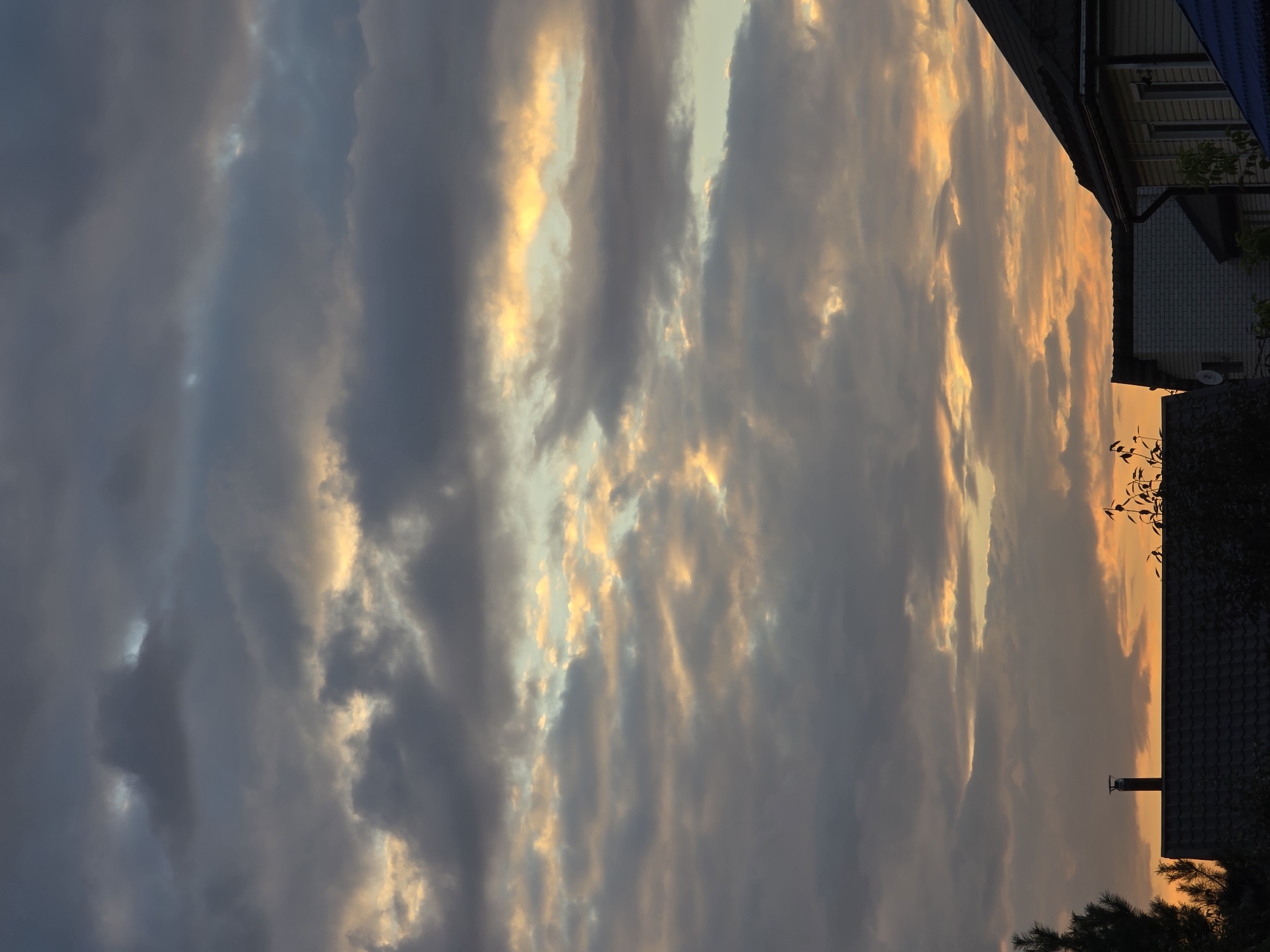 The image shows a cloudy sky with the sun setting or rising, casting warm orange and yellow hues through gaps in the dark grey clouds. The lower portion of the image features the silhouetted rooftops of several houses, indicating a residential area. A tall, dark chimney stands prominently on one of the roofs. The scene appears to be a quiet evening or early morning in a suburban or rural setting, possibly in or near the city of Речицы, Russia. There are no people or active events depicted.