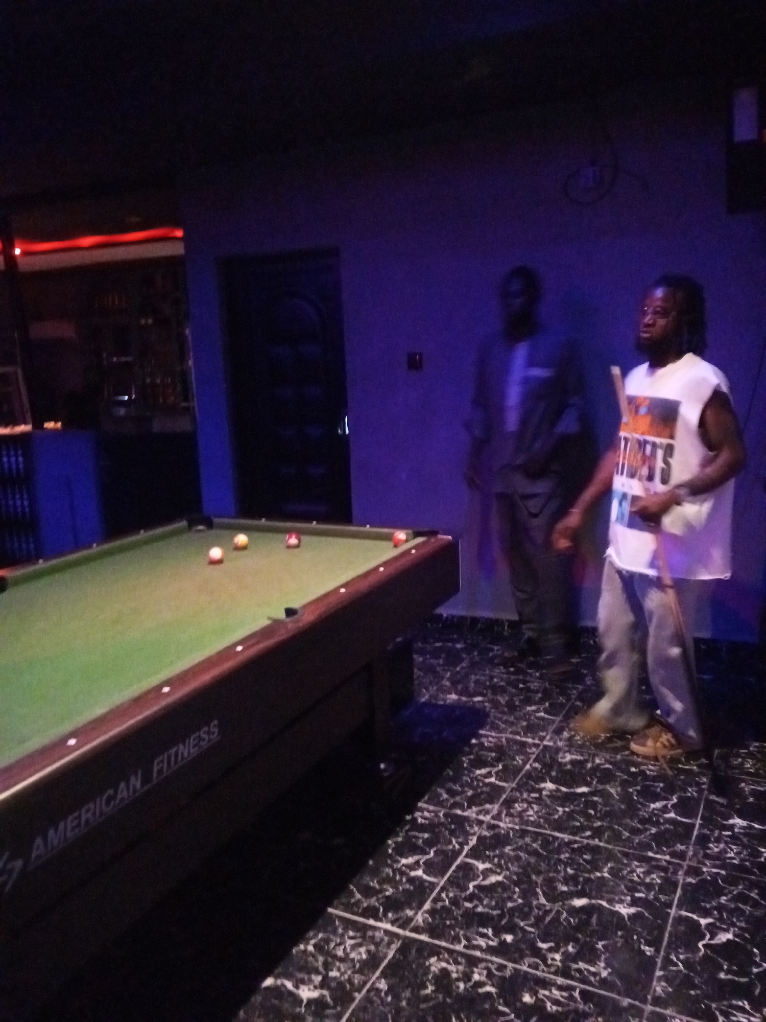 Two men are present in a dimly lit room, likely a bar or recreation center, with a billiard table in the foreground. The man on the right, wearing a white sleeveless shirt with a graphic print and grey pants, is holding a pool cue and appears to be in the process of playing. He has glasses on and dreadlocks. To his left and slightly behind him stands another man in a dark shirt and pants. The billiard table is green with several balls on it, including a red ball, a solid yellow ball, and a striped ball. The room is decorated with dark, marble-patterned flooring and purple lighting on the walls. The overall ambiance suggests a casual social setting. The presence of a billiard table and the attire of the individuals point to a recreational activity taking place. The location is specified as the city of Lokoja, Nigeria.