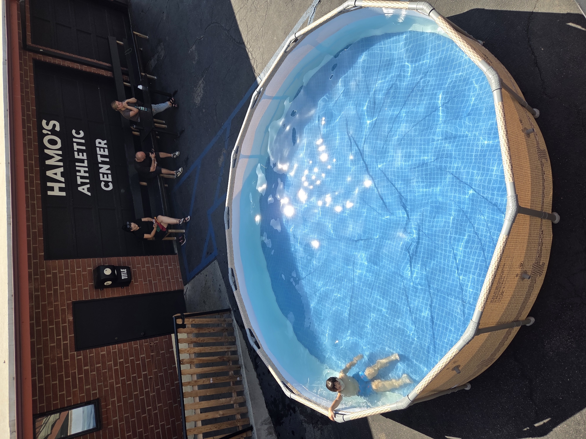 This is a high-angle, medium shot of a young boy in blue swim trunks floating in a round, above-ground swimming pool. The pool is filled with clear blue water, reflecting sunlight, and has a patterned liner. The pool is set on asphalt pavement, possibly in a parking lot, with a shadow falling across it from the right. In the background, there is a brick building with a large black garage door that has white lettering reading "HAMO'S ATHLETIC CENTER". To the left of the garage door, there is a black door and a mailbox with "TITLE" written on it. Several people are seated on a long, dark bench in front of the building. One woman is looking at her phone, another man is looking down at his phone, and a third woman is looking forward with her hands clasped. The location is Glendale, California, United States of America.