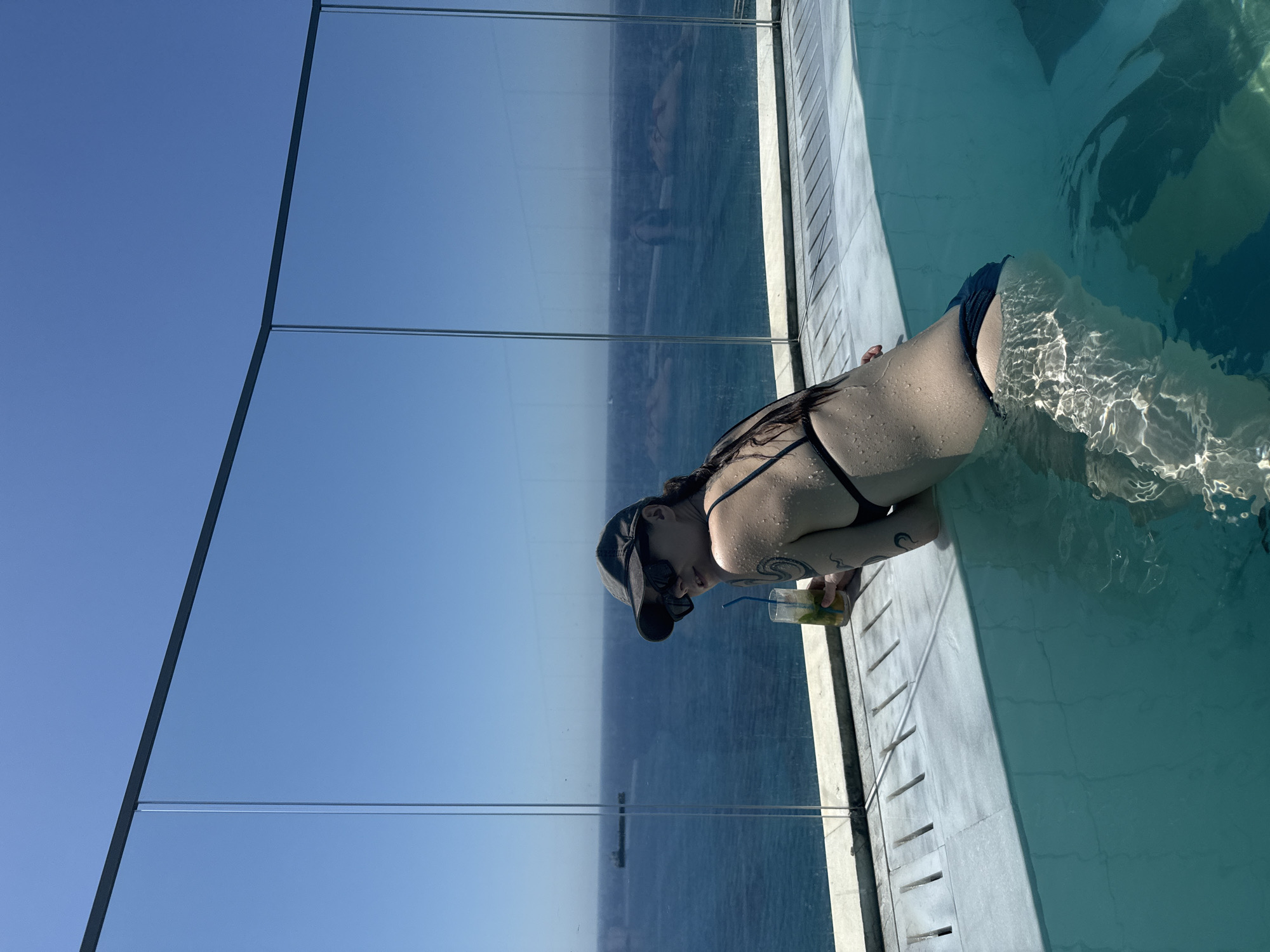 The image depicts a woman in a bikini, wearing a baseball cap and sunglasses, posing in a swimming pool with a glass barrier overlooking the sea. She is holding a drink with a straw. A ship is visible in the distance on the water. The pool appears to be on a rooftop or a high vantage point, with the city of Limassol, Cyprus, potentially visible in the background, given the context of the location. The lighting suggests a sunny day.