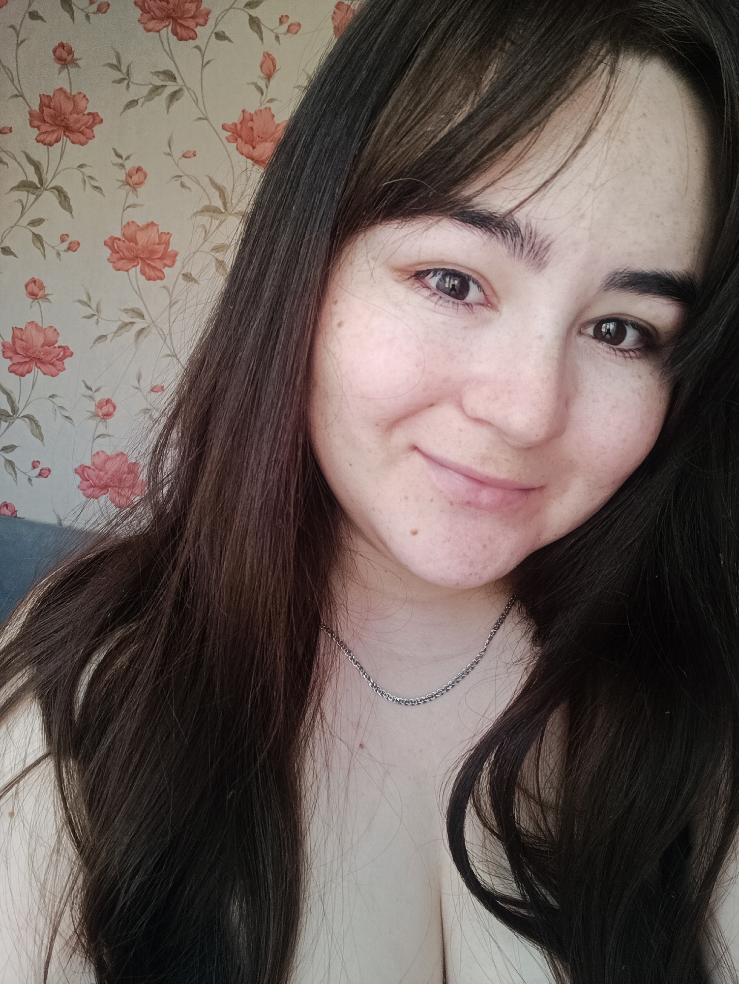 A young woman with long, dark brown hair and prominent freckles on her face is captured in a close-up, chest-up portrait. She gazes directly at the camera with warm brown eyes and a gentle, natural smile. Her hair cascades over her shoulders and chest, partly obscuring her left side. She wears a thin, silver-colored chain necklace.
The background features floral wallpaper with a repeating pattern of reddish-pink flowers and green leaves on a light, possibly cream-colored, ground. A small section of a blue object, likely a piece of furniture, is visible in the lower-left corner, behind her. The indoor setting suggests a personal space, possibly a home in Noviy, Russia. The lighting is soft and even, indicative of daytime. No text is visible in the image.