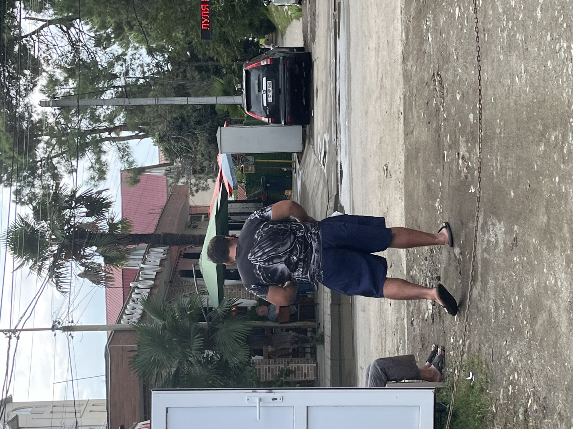 In Ureki, Georgia, a man in a dark t-shirt with a praying hands graphic and dark shorts stands on a dirt and gravel surface. He is facing away from the camera. To his left, a person in gray pants sits on a bench. In the background, a black Honda CR-V SUV is parked, displaying a license plate with the characters "37 NP 066". A building with a red roof is visible behind the SUV, and several trees, including palm trees, are present in the scene. A white metal structure, possibly a kiosk or booth, is situated near the SUV. Red neon lights spelling out Cyrillic text are visible in the upper right portion of the image.