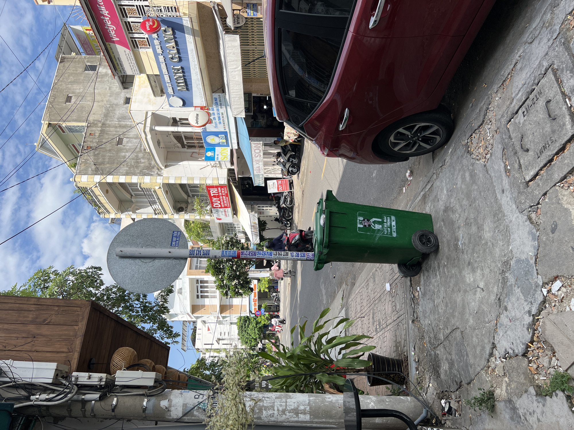 The image captures a street scene in Nha Trang, Vietnam, on a sunny day with a partly cloudy sky. In the foreground, a large green trash bin with wheels stands on the sidewalk, partially obscuring a cracked concrete pavement littered with debris, including cigarette butts. To the right, the rear section of a dark red car is visible, parked along the street. The sidewalk continues to the left, with a potted plant and a table next to a utility pole that is heavily laden with electrical wires and conduits. Further down the street, traffic, primarily motorcycles, is moving along the asphalt road, with several buildings lining both sides. These buildings are of varying architectural styles and heights, many featuring balconies and signs for businesses, some in Vietnamese. A prominent sign for "Jetstar" and another reading "MINH GIANG" are visible. The overall impression is of a typical urban street in Vietnam, with a mix of residential and commercial properties and the constant presence of motorbikes.