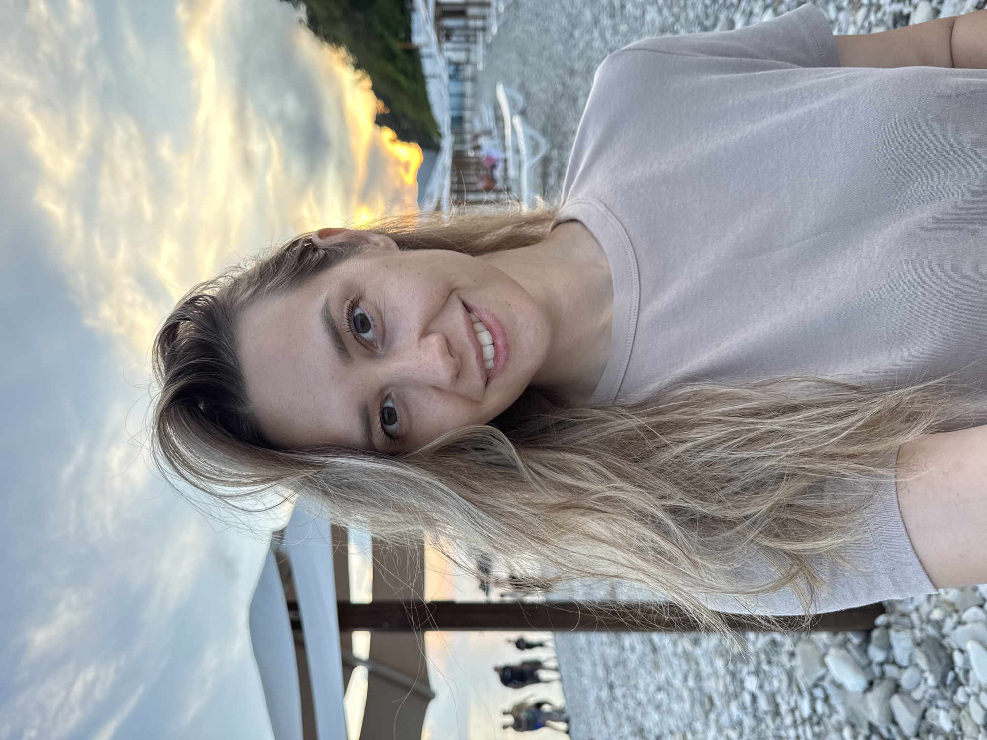 A woman is taking a selfie on a beach in Nebug, Russia. She has blonde hair and is wearing a light-colored t-shirt. The beach is made of pebbles, and there are lounge chairs and umbrellas visible in the background. The sun is setting, casting a warm glow over the scene. Several people are walking on the beach in the distance.