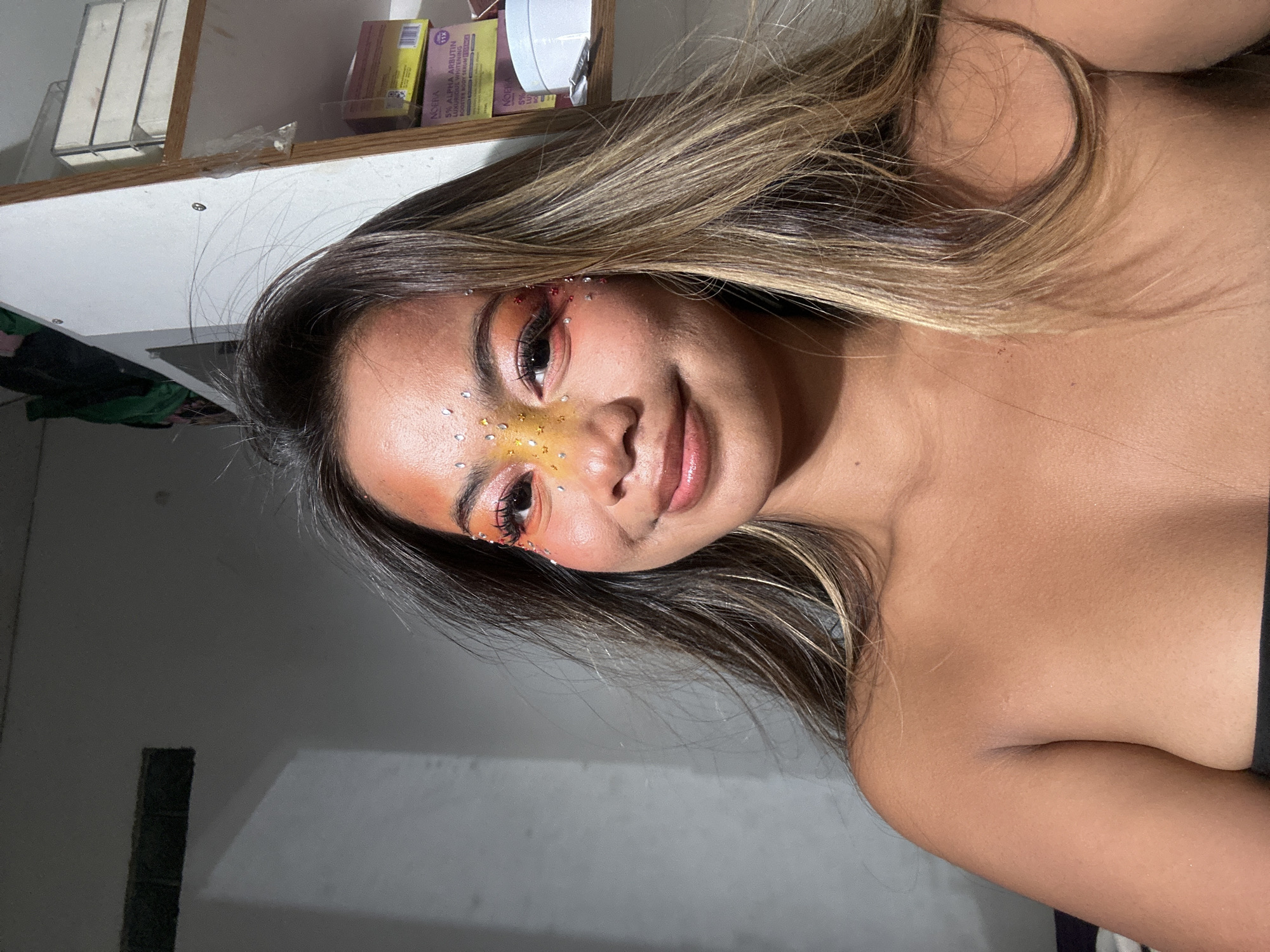 A medium shot of a young woman with brown and blonde highlighted hair, wearing orange and pink makeup with iridescent and gold embellishments on her face. She is smiling slightly and looking at the camera. The background includes a white wall, a shelf with various items on it, and a dark rectangle shape. The location is plausibly indoors. There is no information in the image to determine the city of North Jakarta, Indonesia.