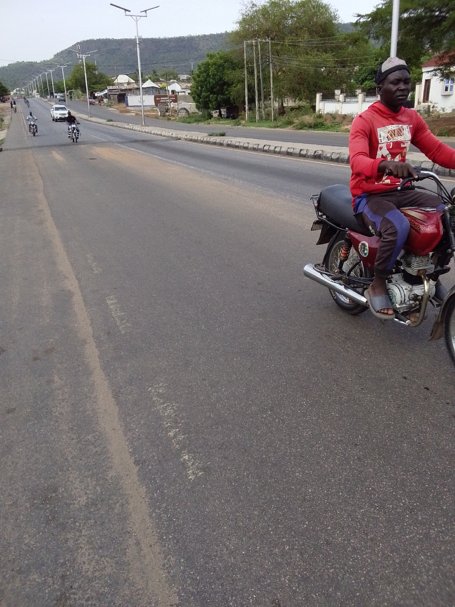 The image shows a man riding a red and black motorcycle on a paved road in Adankolo, Nigeria. The man is wearing a red long-sleeved shirt, dark pants, and sandals. He has a dark complexion and is wearing a cap. The road is wide and stretches into the distance, with other motorcycles and a white car visible further down. There are utility poles lining the side of the road, along with buildings and trees. In the background, a hill or mountain is visible. The sky appears overcast.