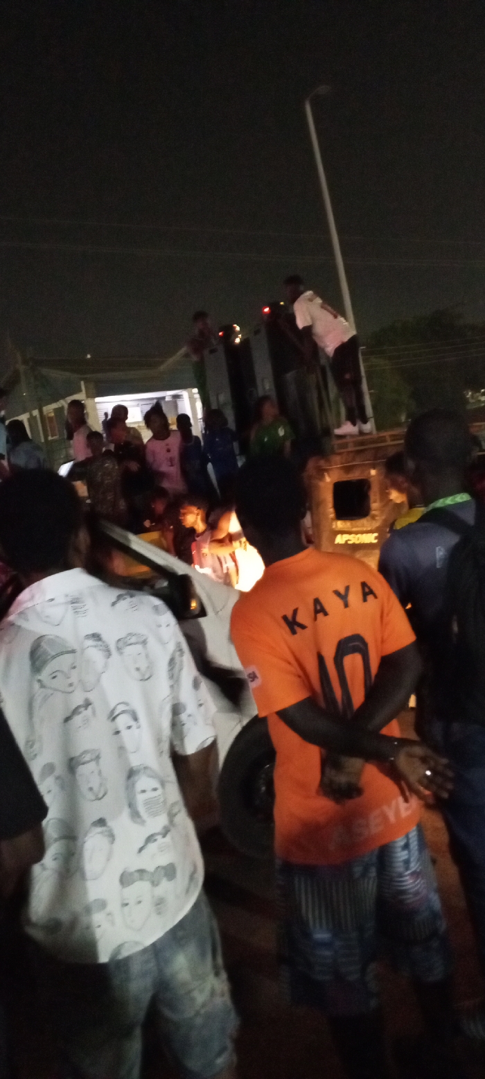 In a dimly lit outdoor scene, a crowd gathers around a truck, some standing atop it. The gathering appears festive, with individuals wearing colorful clothing. One person wears an orange shirt with "KAYA 10" printed on the back, while another sports a white shirt adorned with faces. A street light looms in the background, illuminating the event. The crowd's attention seems focused on the truck.