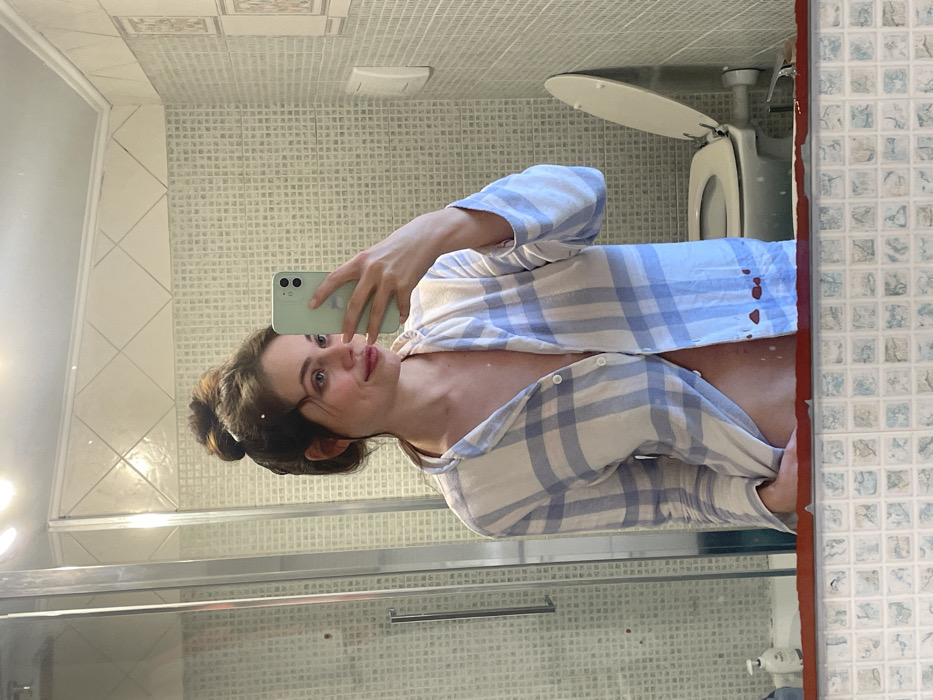 A female subject is depicted in a bathroom, captured via a mirror selfie. The subject, with dark hair tied in a bun, wears a light blue and white plaid button-up shirt, partially unbuttoned to expose her midriff. She holds a light green smartphone in her right hand, using it to photograph her reflection. Her head is slightly turned to her right, and she exhibits a faint smile. The bathroom features light-colored, small square wall tiles, with a decorative border of larger, diagonally set tiles near the ceiling. A glass-paneled shower enclosure is visible on the left side of the reflection, and a white toilet with its seat and lid raised is visible on the right. A white wall-mounted control panel is present above the toilet. A white dispenser bottle is positioned on a surface near the bottom left of the reflection. The scene is located within a private residence in the city of Rome, Italy.