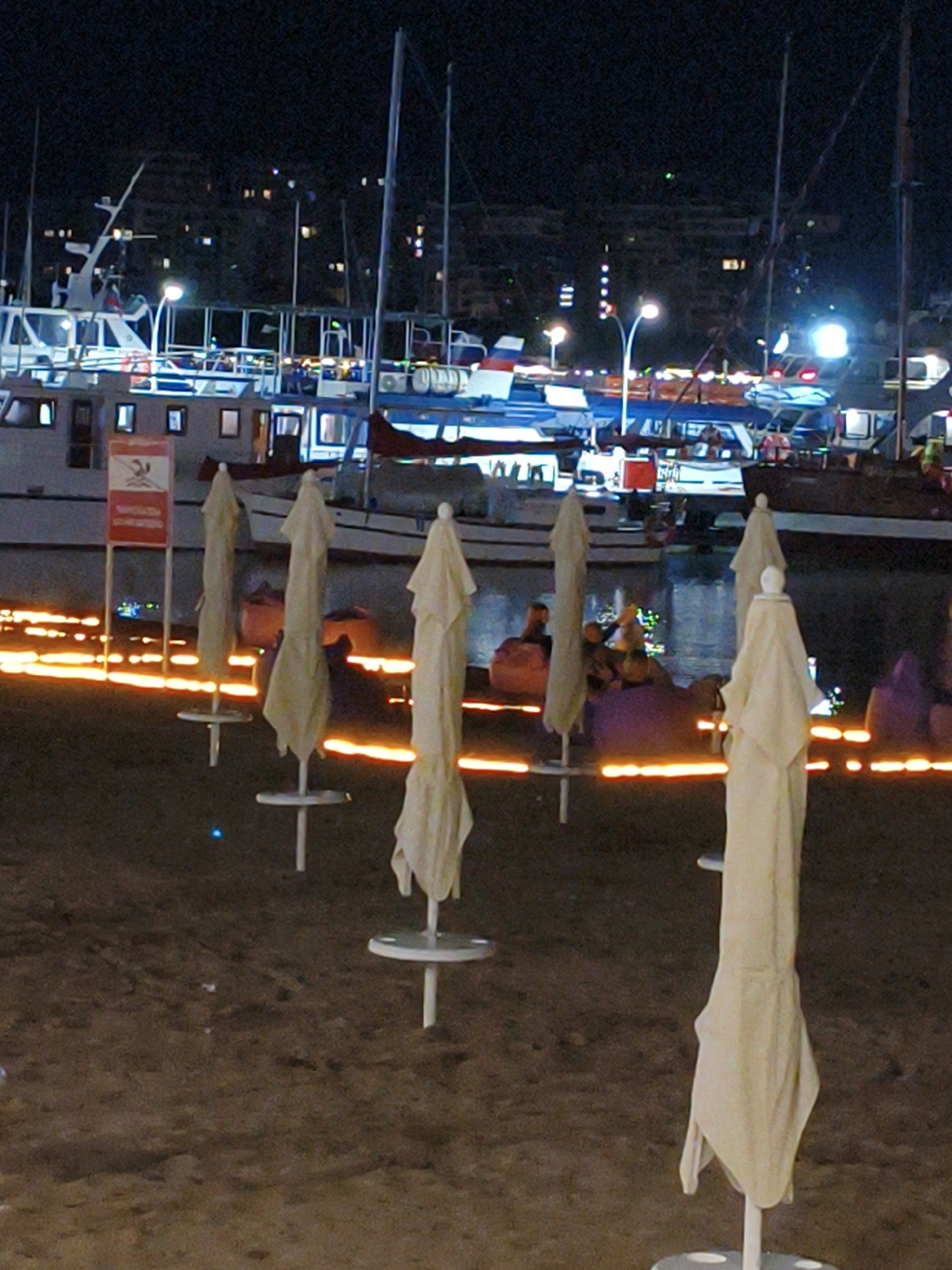 This image depicts a nighttime scene at a waterfront. In the foreground, several closed beach umbrellas stand upright on a sandy surface. Behind the umbrellas, a line of glowing orange lights illuminates the sand near the water's edge.
In the background, several boats are docked in the water. Some of these appear to be larger vessels, possibly ferries or yachts, with illuminated windows and structures. Other smaller boats are also visible. Beyond the boats, a shoreline with buildings and lights suggests an urban or developed area. The overall lighting indicates it is nighttime, with artificial lights from the boats, buildings, and the decorative string lights providing illumination.
There are no people clearly visible in the foreground, but some figures can be discerned in the background, seated on beanbag chairs near the water. The presence of closed umbrellas and the nighttime setting suggest the beach area is not currently in active use for daytime activities, but may be set up for evening ambiance or waiting for dawn. The reflective orange lights create a somewhat festive or romantic atmosphere. A sign with red and white elements is partially visible on a pole in the left foreground, but the text is illegible.