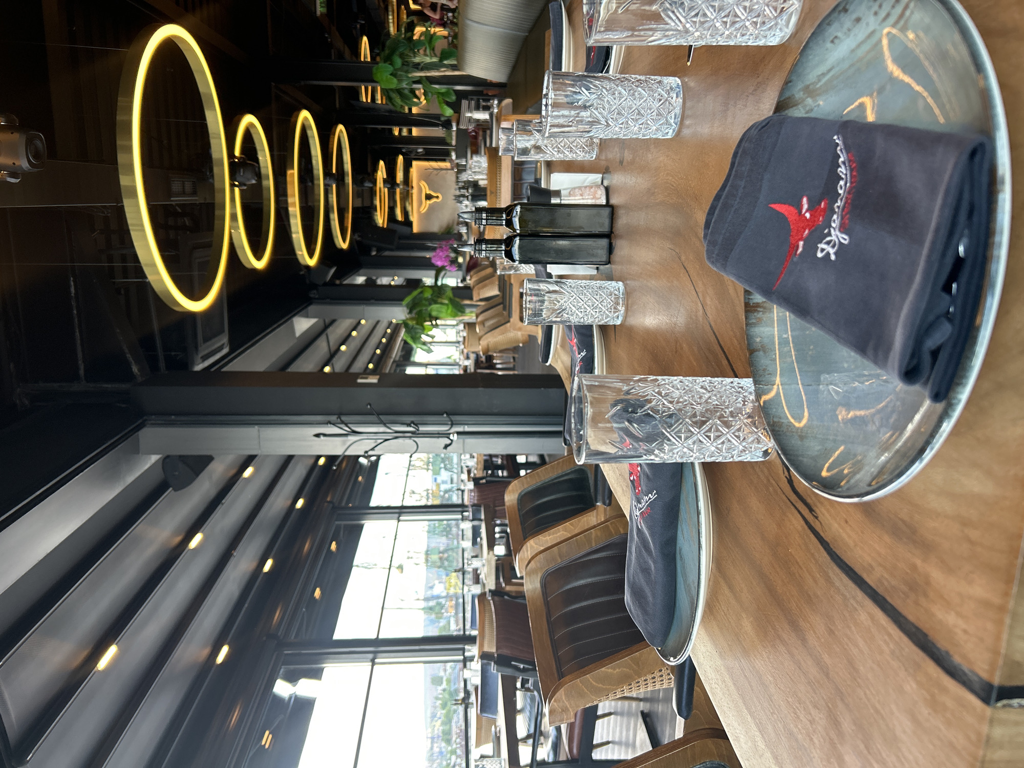 The image shows the interior of a restaurant in Varna, Bulgaria. The restaurant features a long wooden table set for dining, with plates, glasses, and dark-colored napkins with a red bird logo. Two bottles of oil/sauce are placed in the middle of the table. Above the table, there are decorative gold ring-shaped lights. The restaurant has a retractable roof with windows offering an outdoor view of what seems to be a city landscape. There are several wooden chairs along the table. The setting suggests a stylish, possibly upscale dining experience. No people are visible in the image.