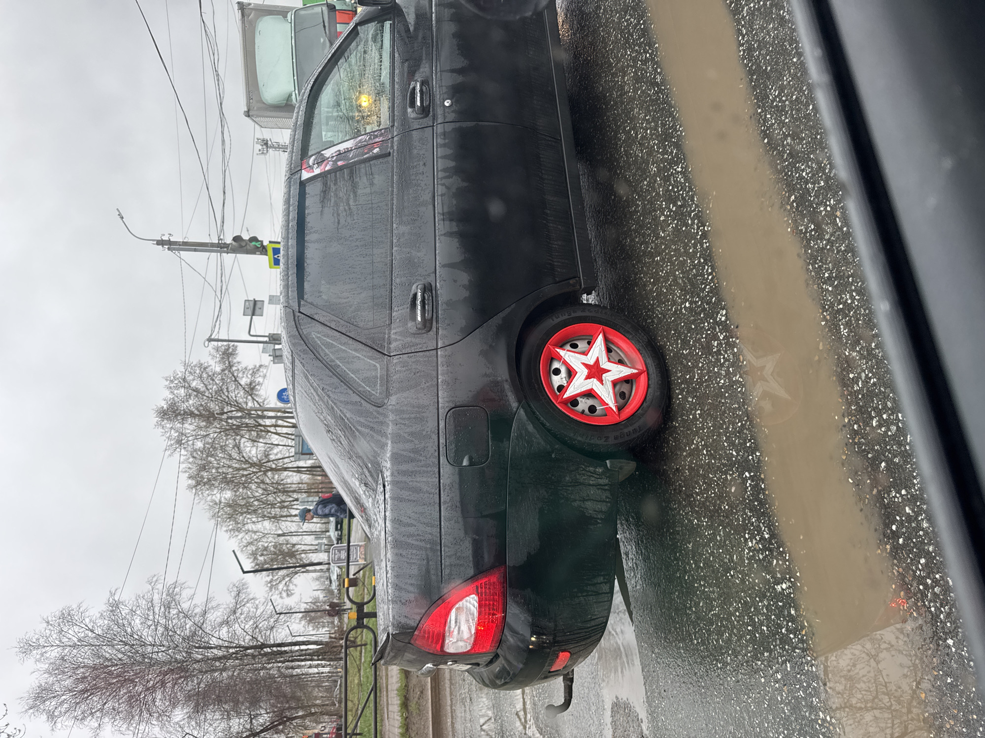 The image depicts a dark-colored Lada sedan parked on a wet street in Chaikovsky, Russia. The car has custom red and white star-shaped wheel covers. The street is wet, with a visible puddle reflecting the car and surrounding environment. In the background, there are trees, power lines, and other vehicles. A pedestrian can be seen in the distance near some railings. The lighting suggests an overcast day.