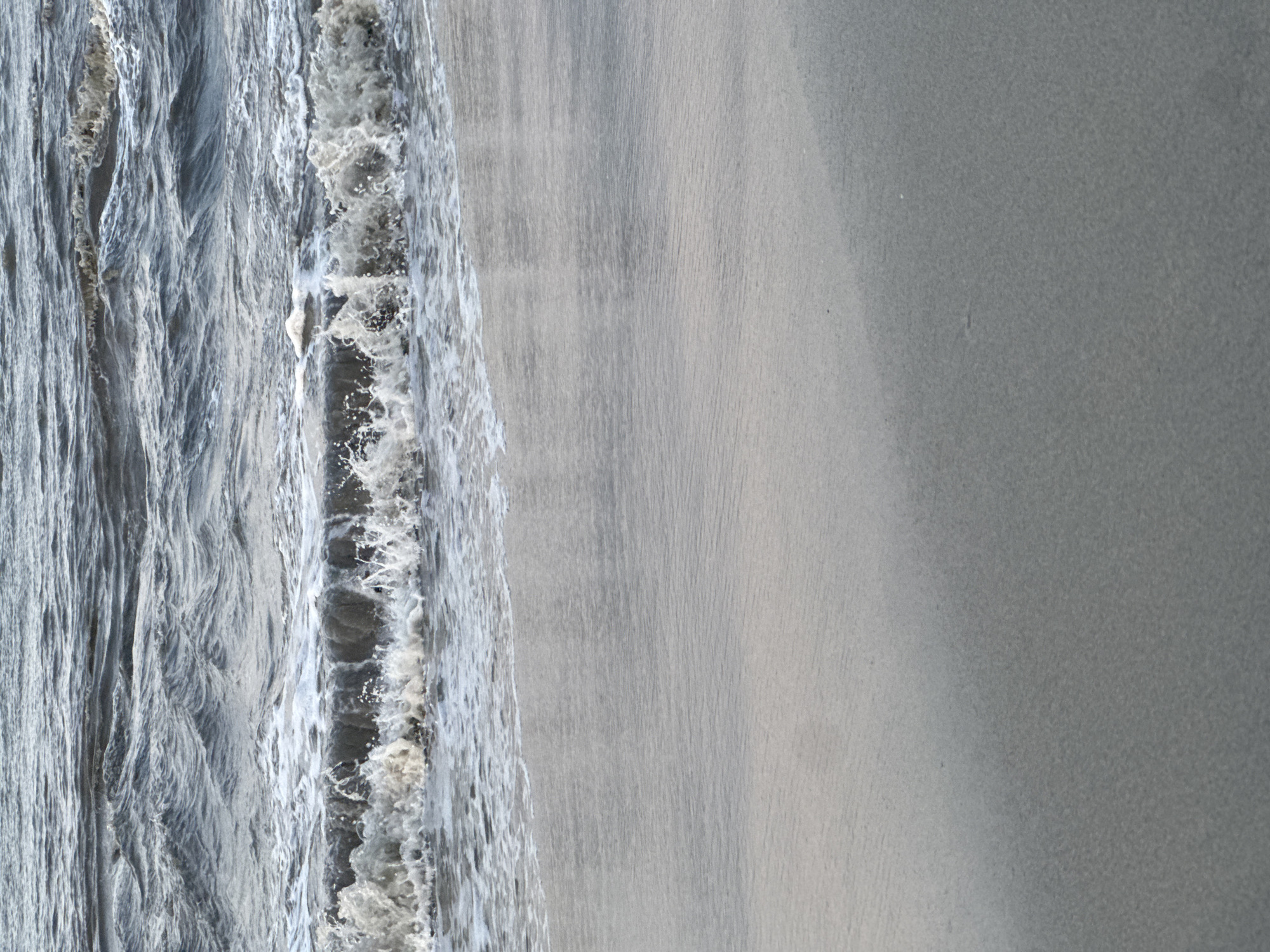 The image depicts a beach scene. The left side shows ocean water with visible waves and foamy spray. The right side exhibits a smooth, sandy beach. The image is rotated 90 degrees clockwise. The location might be in the city of Đà Nẵng, Vietnam.