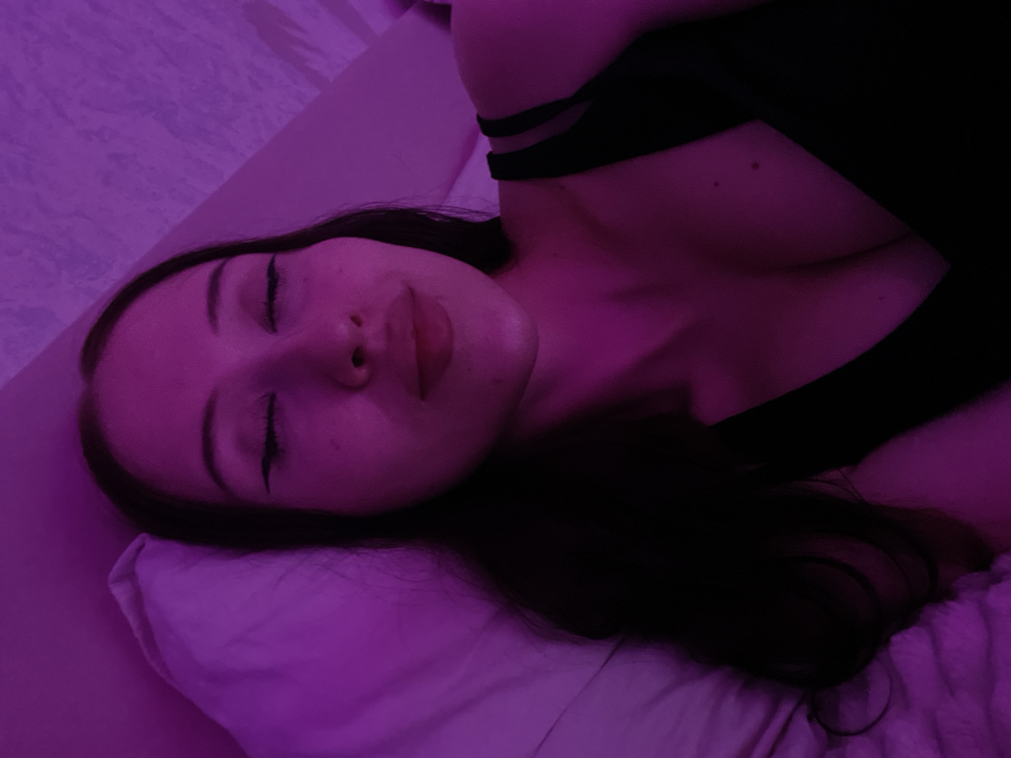 This is a selfie of a young woman lying on a pillow, with her eyes closed and her lips slightly pursed. She has long, dark hair and is wearing a black top. The scene is illuminated with purple lighting. The background appears to be a textured wall and bedding. There is no specific information available about the location in Votkinsk, Russia.