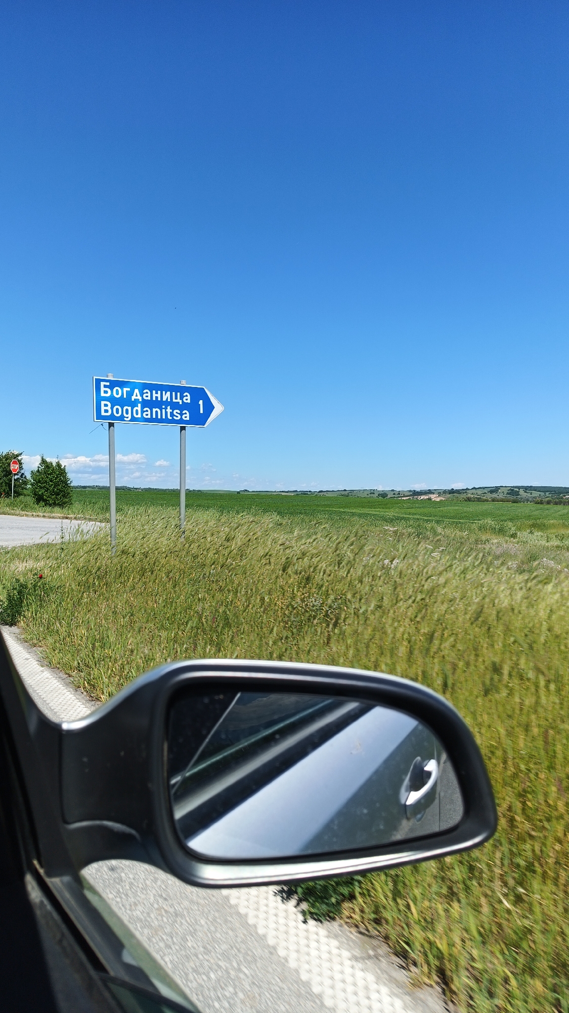 The image shows a road sign indicating the direction to Bogdanitsa, Bulgaria. The sign is blue with white lettering in both Cyrillic and Latin alphabets, displaying "Богданица Bogdanitsa" and a number "1" with an arrow pointing to the right. The sign is located at a road intersection in a rural area, with a field of green and yellow grass stretching to the horizon. To the left of the sign is a stop sign and some greenery. The image is taken from inside a car, with a side-view mirror visible in the foreground. The mirror reflects the car's door handle and a glimpse of the passing scenery. The sky above is clear and blue.