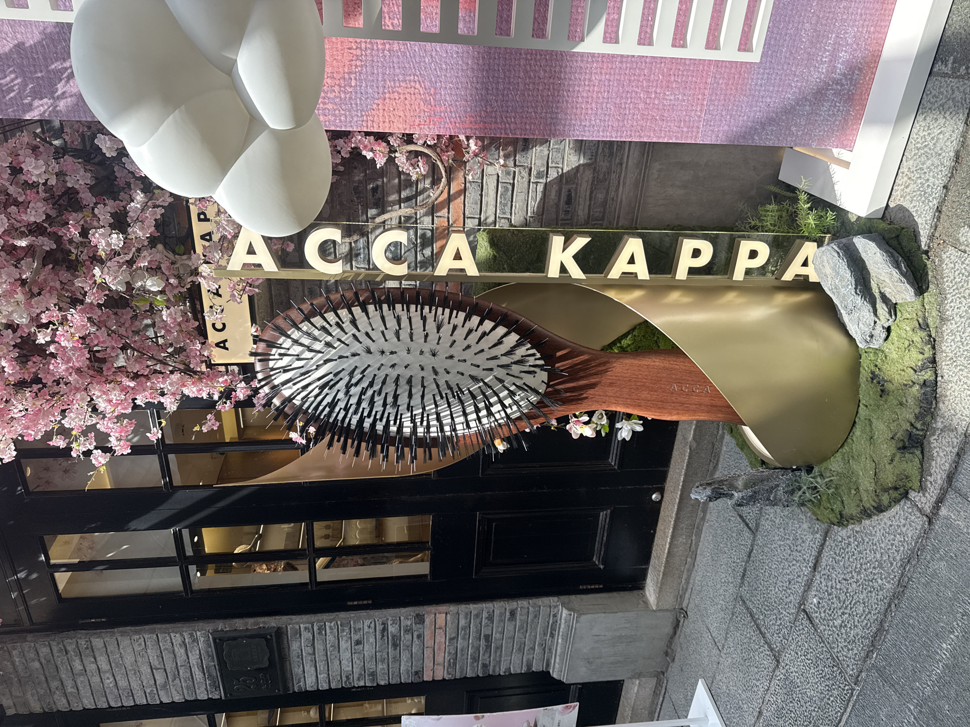 The image shows a storefront in Shanghai, China, featuring a large decorative hairbrush as part of a display. The storefront has a brick facade and a black door with multiple glass panes. Pink artificial cherry blossoms hang from a structure in front of the building. To the right of the hairbrush, large, white cloud-like sculptures and a pink and purple patterned panel are visible. Vertical gold lettering spelling "ACCA KAPPA" is integrated into the display. The ground is paved with grey cobblestones. There are no people visible in the image.