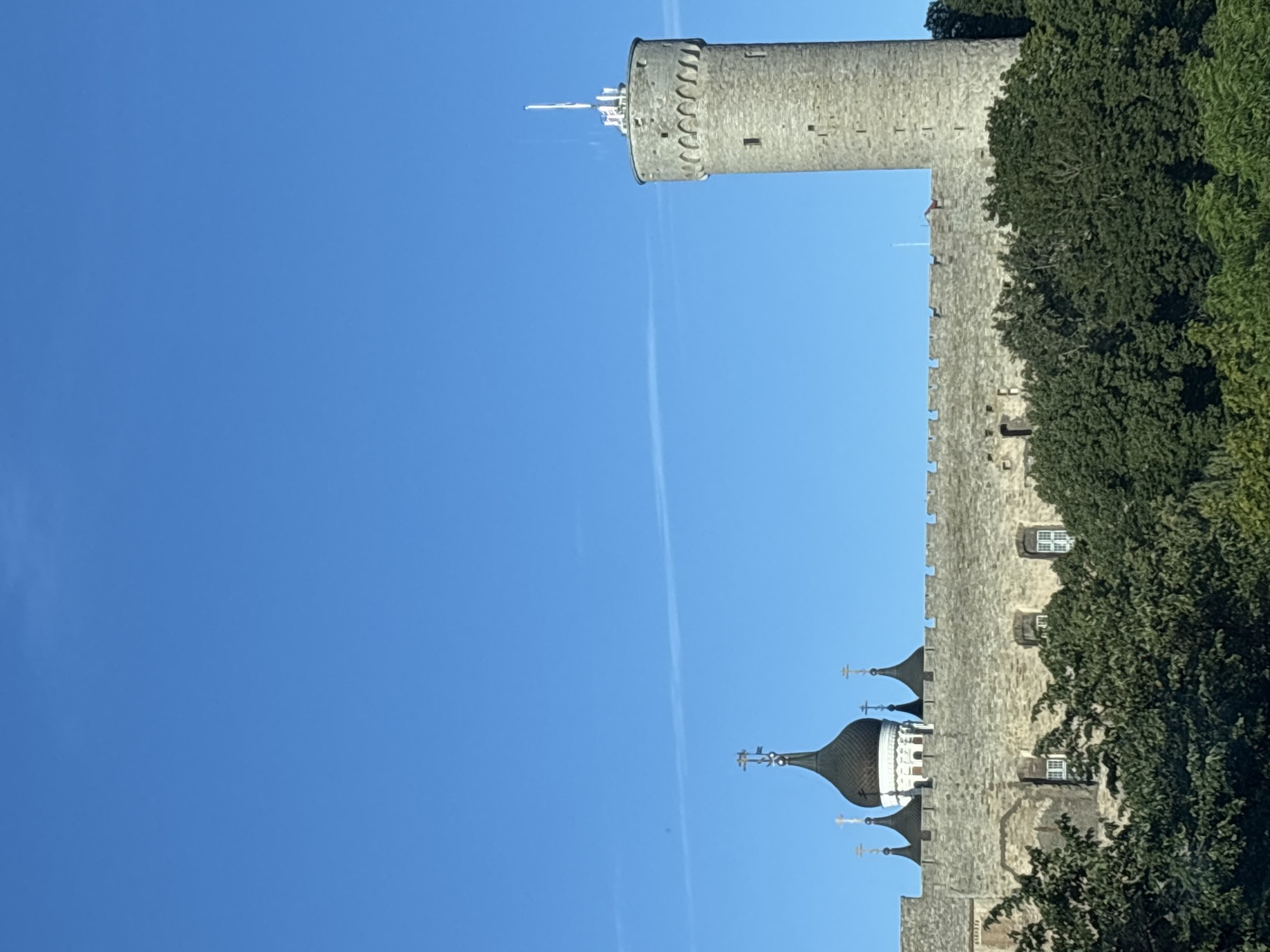 The image depicts the Toompea Castle complex in Tallinn, Estonia, set against a clear blue sky. The castle features a stone wall with crenellations, behind which several dark, onion-domed church spires with crosses are visible. To the right of the castle wall, a tall, cylindrical stone tower with arched windows near the top stands prominently. A flag on a pole is positioned at the very top of this tower. Lush green trees form a dense layer in the foreground, partially obscuring the lower sections of the castle wall. The scene appears to be captured during daylight, with the sun illuminating the stone structures and foliage.
