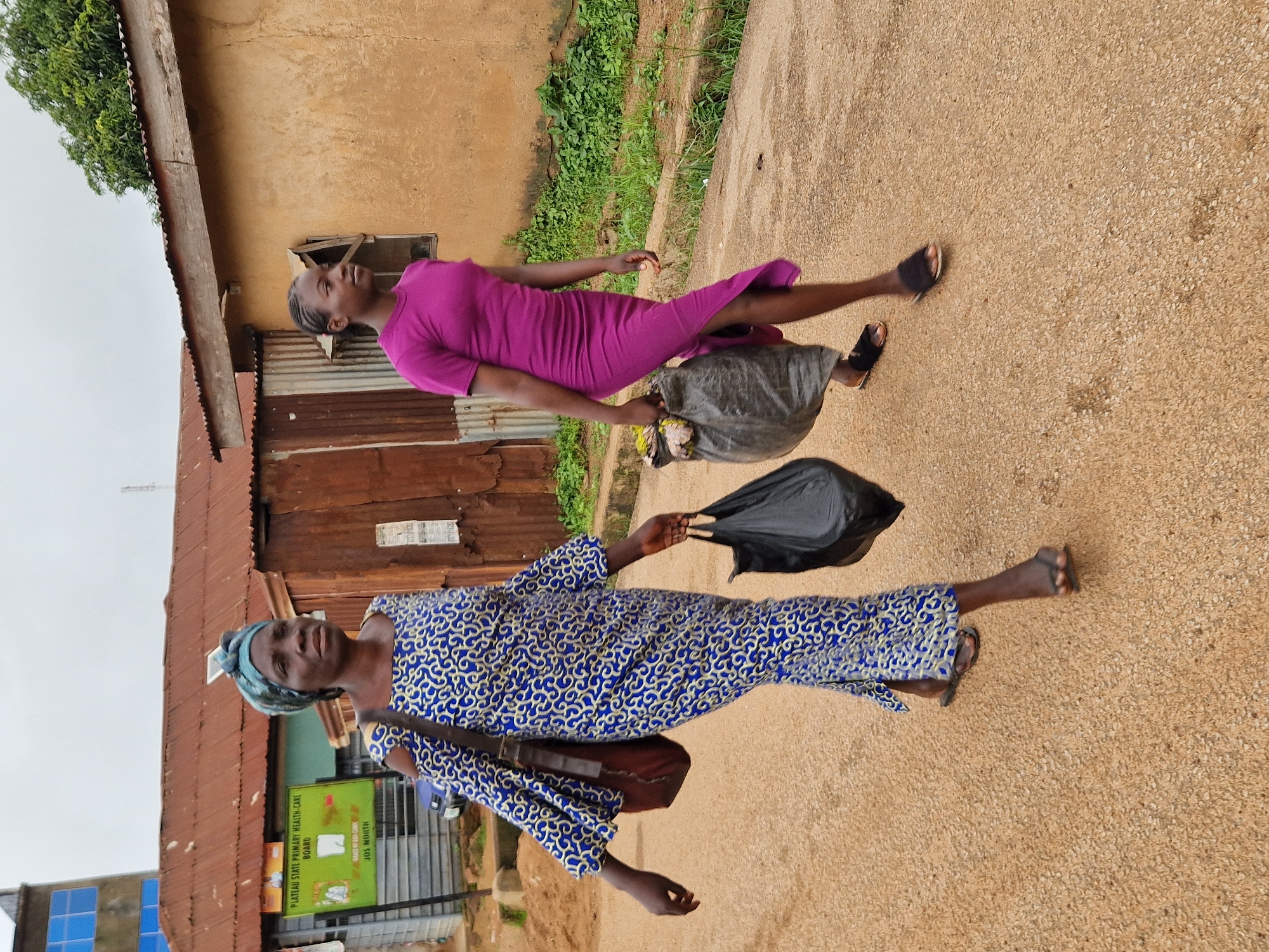In Kabong, Nigeria, two women walk along a dusty road. The woman in the foreground on the left, wearing a blue and white patterned dress and a blue headscarf, carries a black plastic bag in her right hand and a brown shoulder bag across her left shoulder. The woman on the right, in a purple knee-length dress, walks behind and to the right of the first woman, carrying a large, dark plastic bag filled with items in her right hand. The background features a building with a corrugated metal roof, a sign that reads "PLATEAU STATE PRIMARY HEALTH-CARE BOARD," and a smaller, ochre-colored building with a narrow window. Overcast skies suggest a cloudy day.