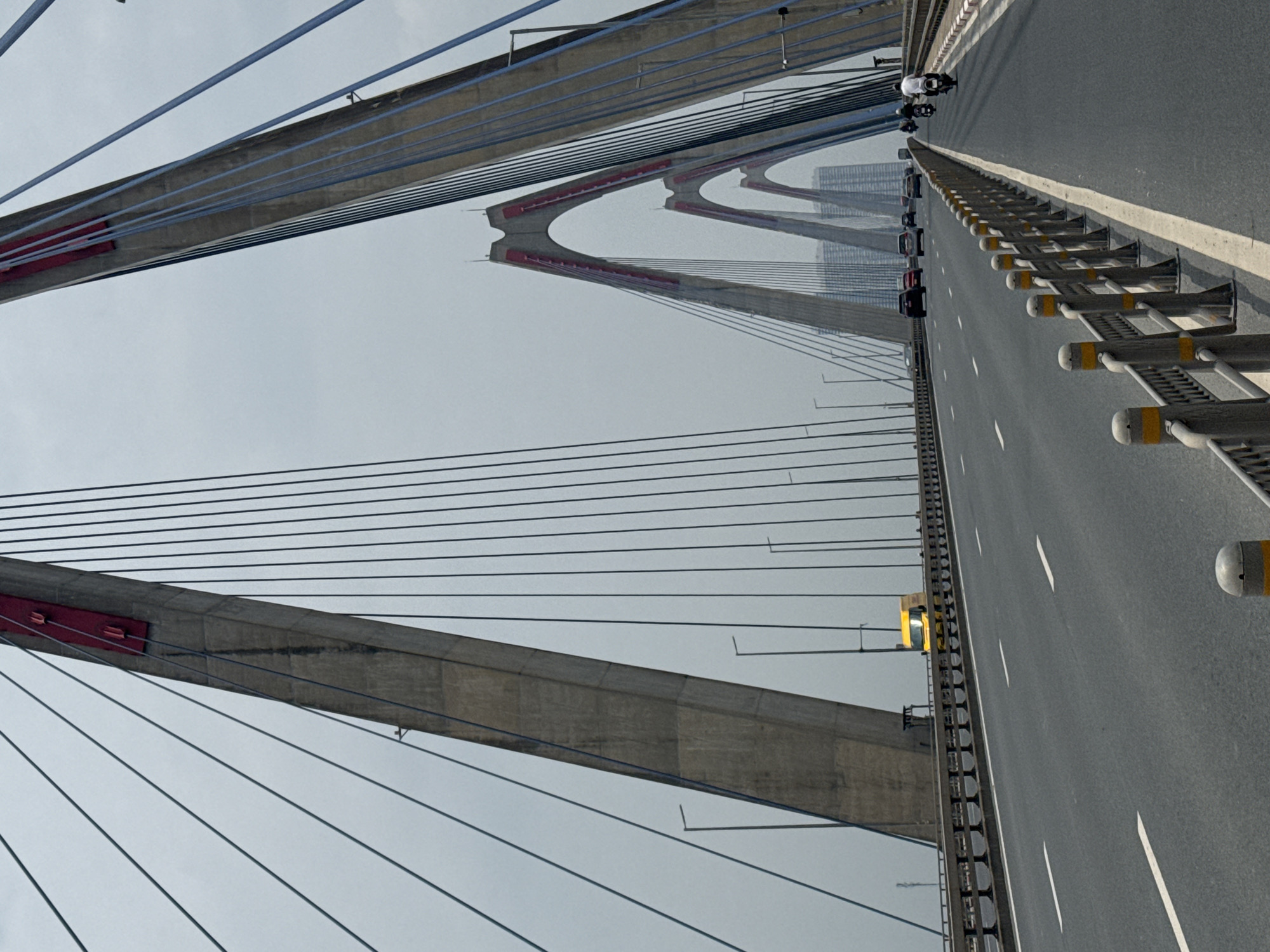 The image depicts a view of the Nhật Tân Bridge in Hanoi, Vietnam. The bridge is a cable-stayed structure characterized by its large, concrete pylons that arch upwards and are adorned with red accents. A multi-lane highway runs across the bridge, with traffic including cars, a yellow truck, and motorcycles. The sky is overcast, and the surrounding environment appears to be an urban landscape with buildings visible in the distance. The perspective suggests the image was taken from a moving vehicle on the bridge.