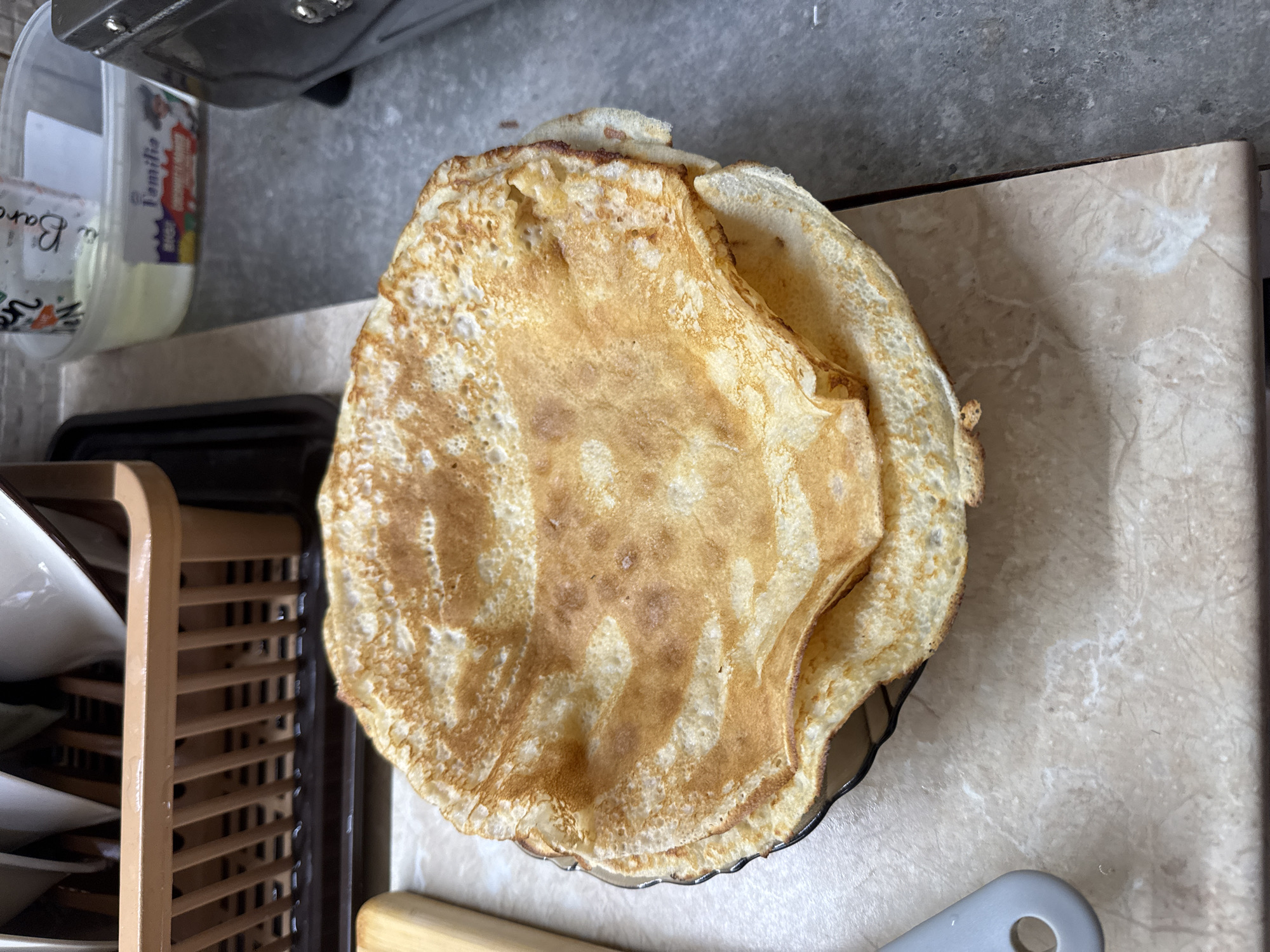 The image displays a close-up, elevated view of a kitchen counter in Unknown, Unknown, featuring a stack of freshly prepared food. The primary subject is a substantial stack of approximately eight to ten thin, circular pancakes or crepes, exhibiting varied golden-brown coloration and a textured surface, consistent with frying. These are arranged on a dark-edged, shallow dish or plate, which is resting on a light-colored, possibly marble-patterned, countertop.
To the upper left, a section of a light brown plastic dish drying rack is visible, holding multiple inverted white ceramic cups or bowls. Adjacent to the pancake stack on the left, a light wooden object, potentially a cutting board or utensil handle, is partially observable. In the lower left corner, a grey plastic utensil handle with a circular hole is also present on the countertop.
The countertop surface transitions from the light-colored, patterned material on the left to a darker, possibly gray or concrete-like, surface on the right. In the upper right background, on the darker counter, a transparent plastic container with a white substance is visible. The container bears partial labels, including the word "Familia" in colored text and other indistinct script. Adjacent to this container, a dark-colored kitchen appliance, possibly a toaster or similar device, is partially discernible on the far right. No individuals are present in the image.
