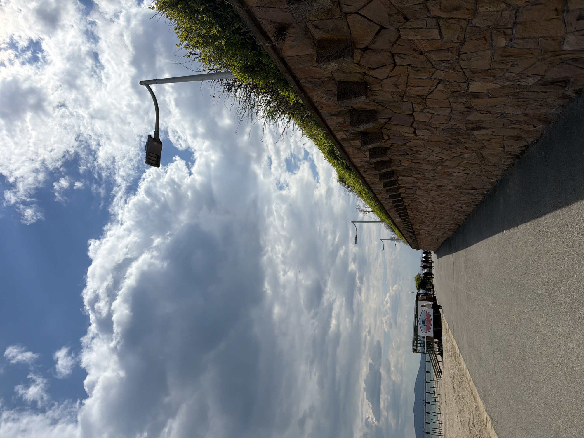The image depicts a promenade in Burgas, Bulgaria. The scene is viewed from a ground-level perspective. A paved path runs along the right side, adjacent to a stone wall topped with greenery. A lamppost is visible to the left, against a backdrop of a cloudy sky. In the distance, structures resembling beachside establishments and human figures can be discerned. A sandy beach and the sea are visible at the horizon line.