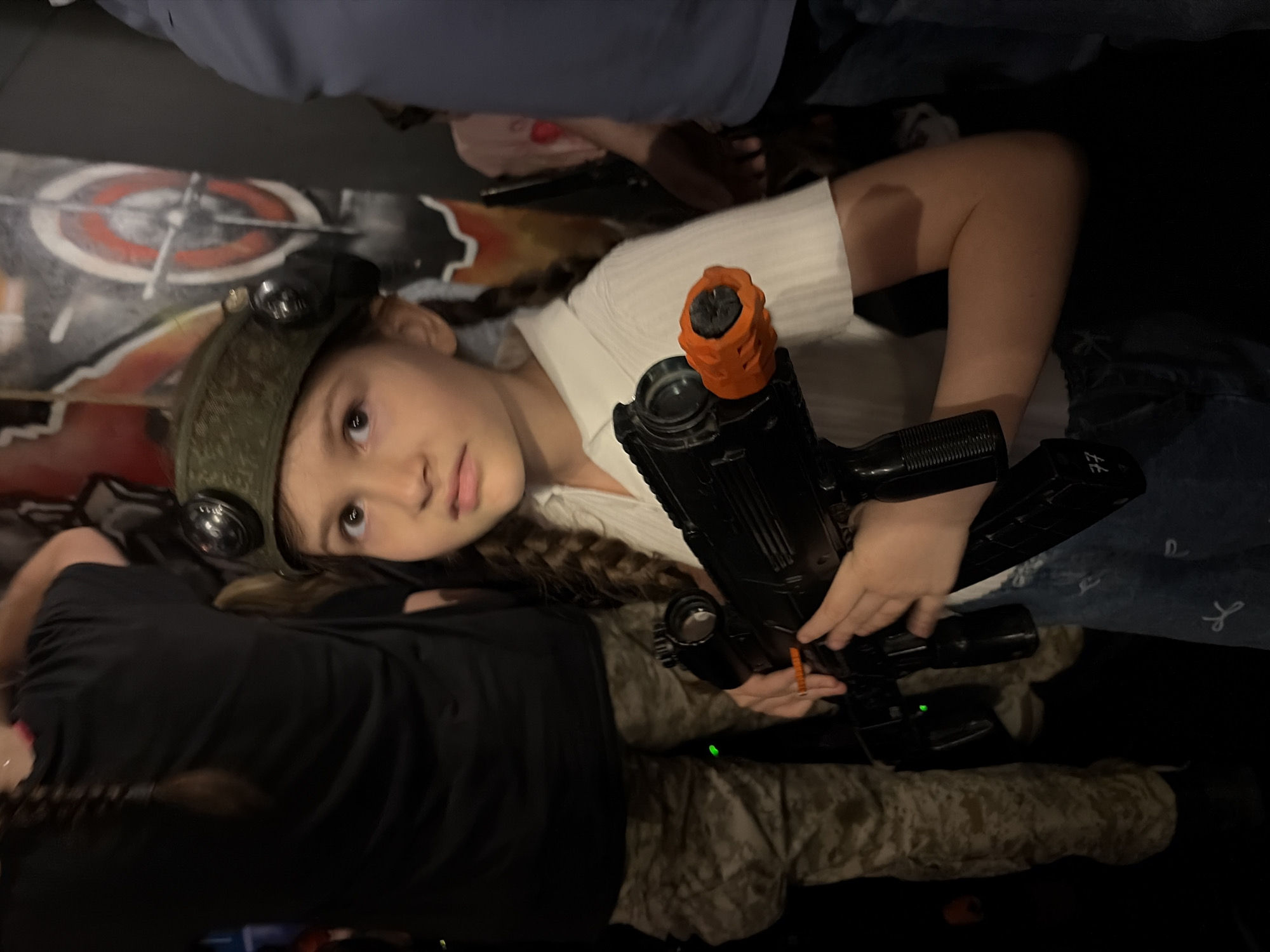 A young girl wearing a camouflage hat with attached lights is holding a black toy gun with an orange tip. She is looking at the camera with a neutral expression. Another person is visible wearing a black shirt and camouflage pants. The background includes a wall with a target design. The location is not identifiable as Moscow, Russia.